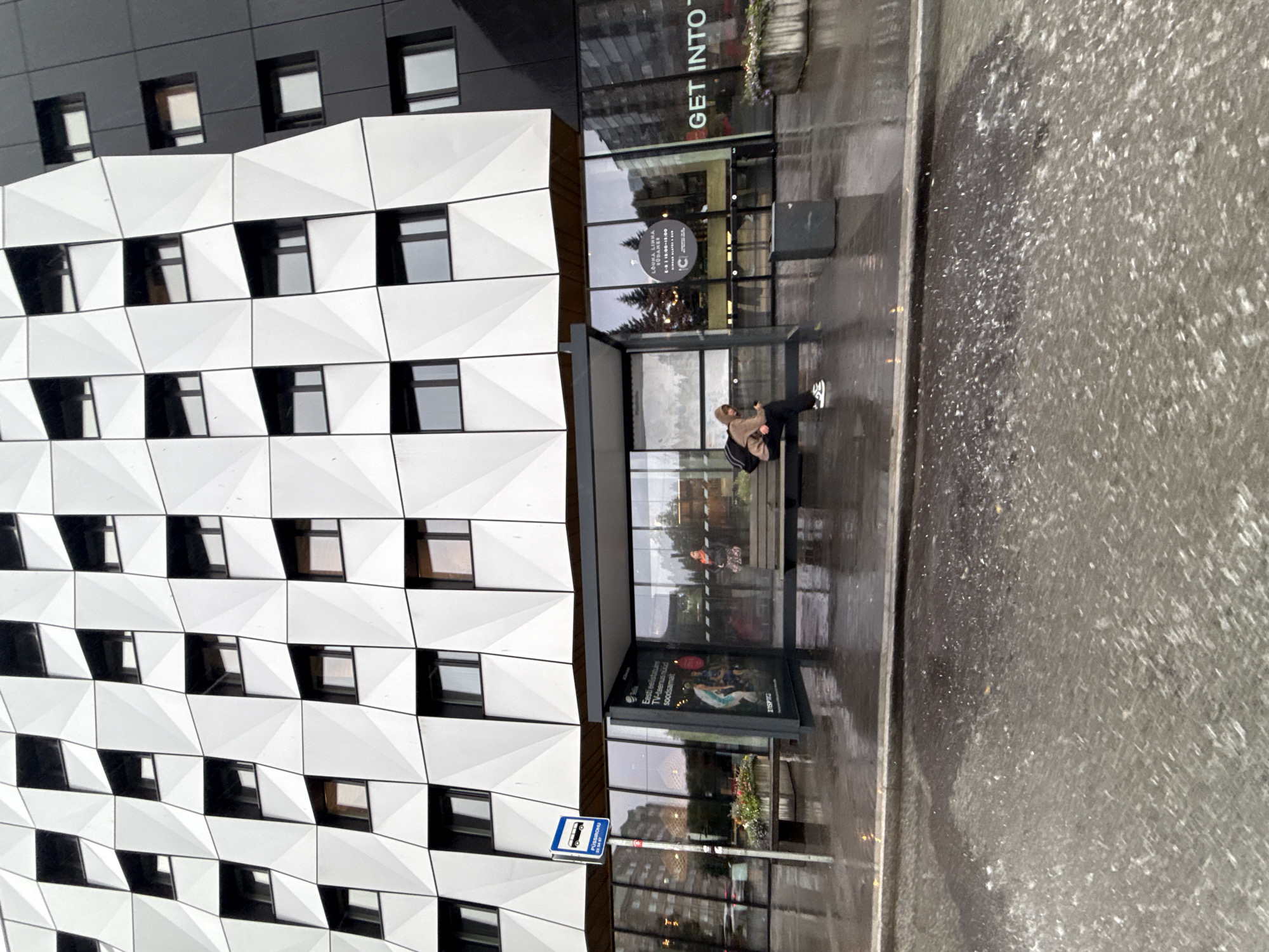 A person sits alone on a bench at a bus stop in front of a modern building with a geometric facade. The street is wet, suggesting it has recently rained or is currently raining, with water reflecting the surrounding lights. A bus stop sign is visible on the left. The building's facade features alternating panels of white geometric shapes and black sections, with numerous windows. The bus shelter has advertising posters displayed on its glass. The overall atmosphere is somewhat subdued due to the weather. This scene is located in Tallinn, Estonia, specifically at a bus stop near the geometric building.