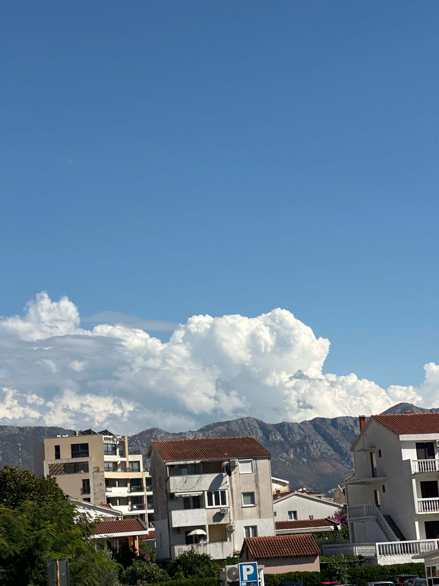 The image depicts a view of buildings and mountains under a bright blue sky with dramatic cumulus clouds in Budva, Montenegro. In the foreground, several residential buildings with red-tiled roofs and white facades are visible. One building on the right features multiple balconies with white railings. Another building on the left is taller and appears to be a hotel, with a sign that reads "HOTEL Z ...". Green trees partially obscure some of the buildings. In the background, rugged, sparsely vegetated mountains rise against the sky, with the large white clouds casting shadows. The overall scene suggests a sunny day in a coastal town with mountainous terrain.