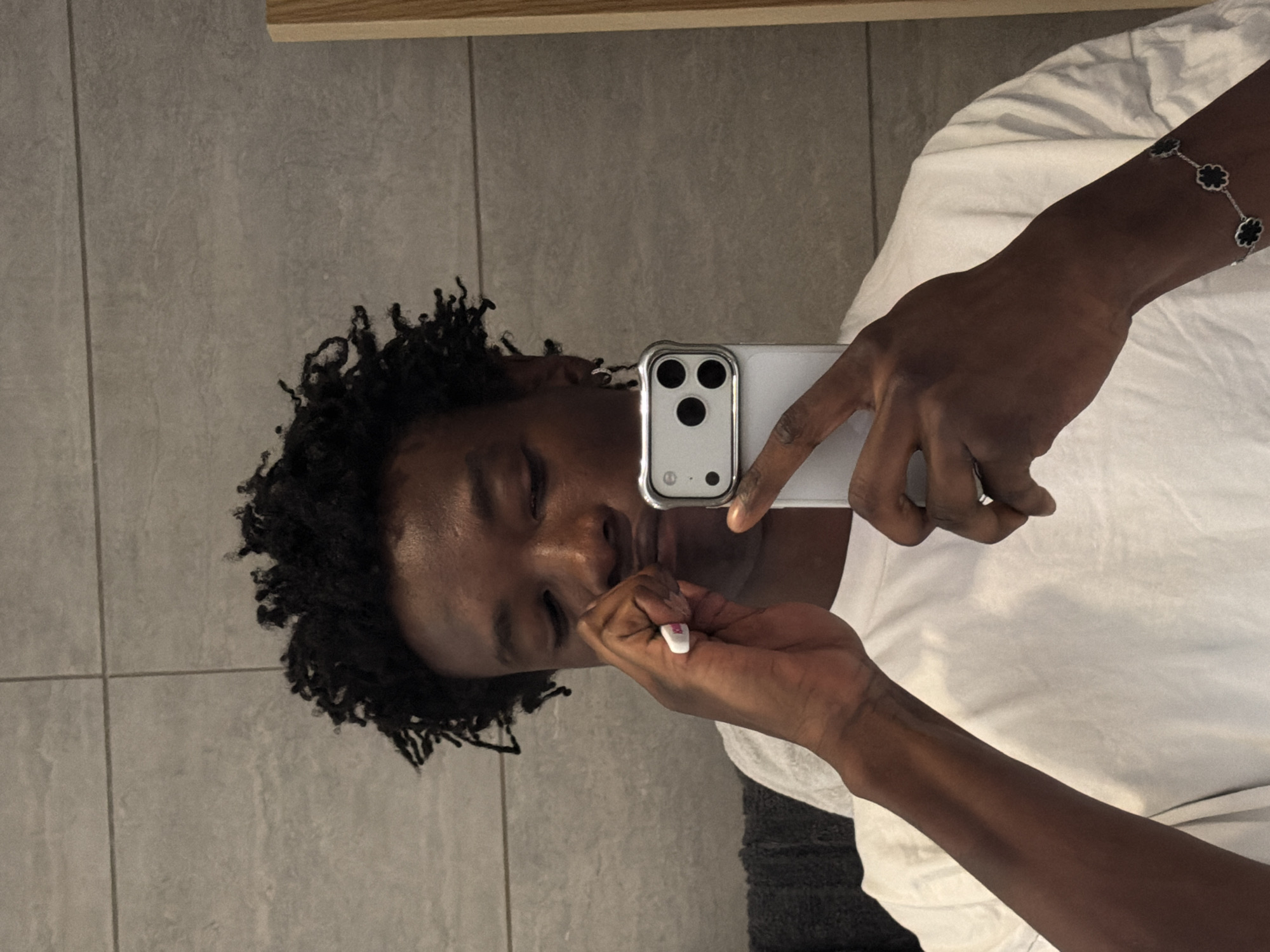 The image shows a close-up, waist-up, selfie-style shot of a young Black male with dark, curly hair. He is wearing a plain white t-shirt and a silver bracelet with black clover-shaped charms on his left wrist. He is holding a white object, possibly a cotton swab, near his mouth with his right hand, and his left hand is partially obscured as he holds a smartphone with a silver case and three black camera lenses. The background appears to be a tiled bathroom wall, with a dark grey towel visible on the left and a wooden door frame on the right. The overall lighting suggests an indoor setting. The image does not contain any elements that can identify its location within the city of London, United Kingdom.