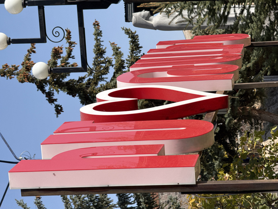 The image displays a large, three-dimensional public sign spelling "ԵՍ ❤️ ԹԱԼԻՆ" (I love Talin) in Armenian script with a central heart symbol. The sign features red frontal surfaces and white side panels, oriented diagonally from the lower left to the upper right, and is supported by dark metal posts. The background consists of a clear blue sky above and numerous evergreen trees, some exhibiting visible cones. To the right of the sign, a black metal lamppost with multiple white spherical light fixtures and decorative scrollwork is present. Further right, a light-colored statue depicting a robed figure with a head covering is visible in profile, partially obscured by foliage. The scene is an outdoor setting illuminated by bright daylight, located in Talin, Armenia. No individuals are present.