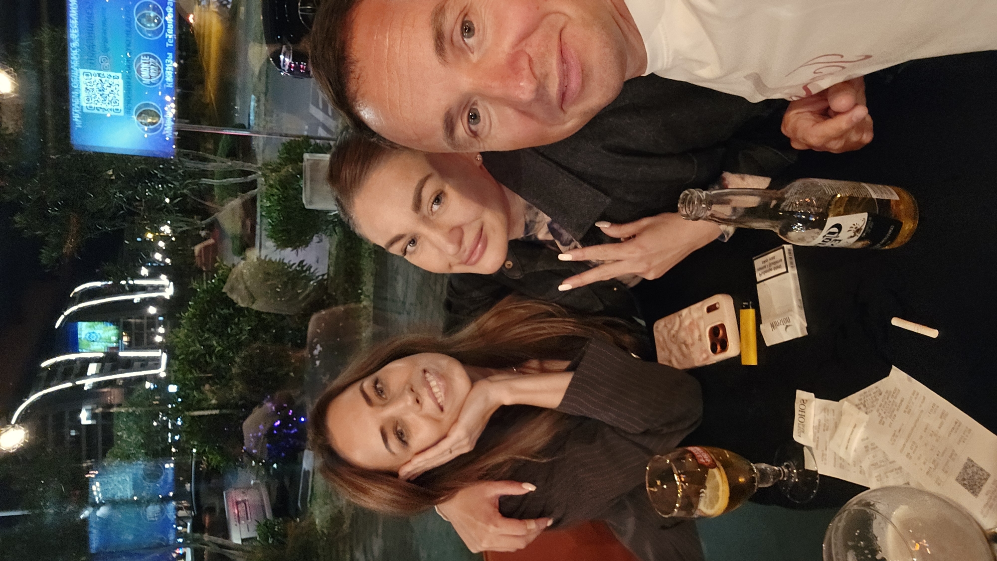 The image depicts three individuals, likely friends or acquaintances, posing for a selfie or photograph at a table in an outdoor setting. A man with a close-cropped haircut appears at the top of the frame. Beneath him, two women are positioned closely together, both smiling. The woman at the bottom rests her chin on her hand, and the other woman holds up two fingers in a "peace" sign.
The table is littered with items: an empty Corona beer bottle, a pack of Marlboro cigarettes, a yellow lighter, a cell phone, a cigarette, a wine glass containing a beverage and lemon wedge, an empty glass, and a crumpled receipt with a QR code.
The background features an urban environment with buildings illuminated at night. A sign displays text in Russian and what seems to be a QR code. Based on the building design and signage visible, it is highly probable the location is in the city of Bar, Montenegro.