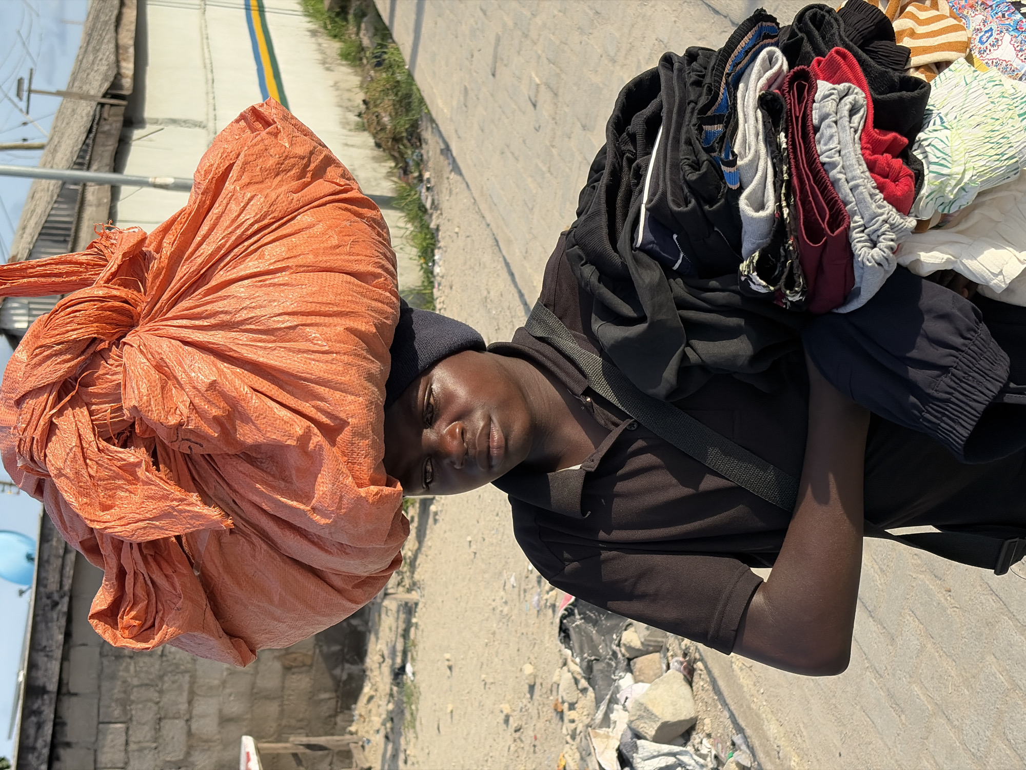 The image shows a man in Igboefon, Nigeria carrying two large bundles. A large orange bag is balanced on his head. He also carries a pile of clothes and other items on his shoulder. He wears a dark beanie, dark collared shirt, and a shoulder strap. The background includes a paved road, dirt, and building structures.