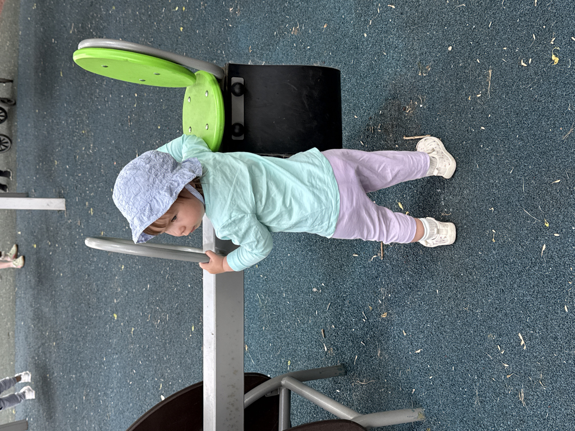 A young child, wearing a light blue sun hat, a light green long-sleeved shirt, and light purple pants, is using playground equipment. The equipment has a green and silver component near the child's head and a black component near the child's midsection. The playground surface is a dark blue rubber material. The image suggests an outdoor location, possibly in Moscow, Russia.