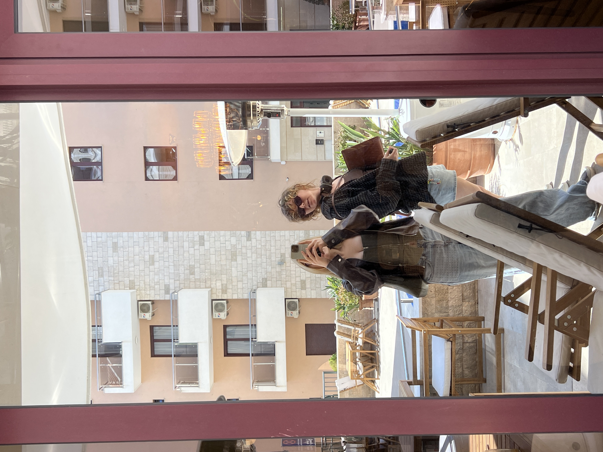The image depicts a reflection of a woman and the exterior of a building in what appears to be Bečići, Montenegro. The reflection is seen through a reddish-brown-framed window, potentially of a restaurant or establishment.
The woman is holding a smartphone as if taking a photograph or selfie. She is wearing sunglasses, a dark jacket or shirt with a plaid pattern, and denim shorts. She has curly blonde hair and is wearing headphones around her neck. She is holding a brown bag or purse.
The reflection also shows the exterior of a multi-story building with light peach-colored walls, dark brown window frames, and several air conditioning units mounted on the facade. A portion of a decorative overhead light fixture is visible. The building appears to be a hotel or apartment complex.
A patio area with wooden chairs and white cushions is also visible in the reflection. A couple of people are seated on one of the lounge chairs. The setting suggests an outdoor space adjacent to a building.