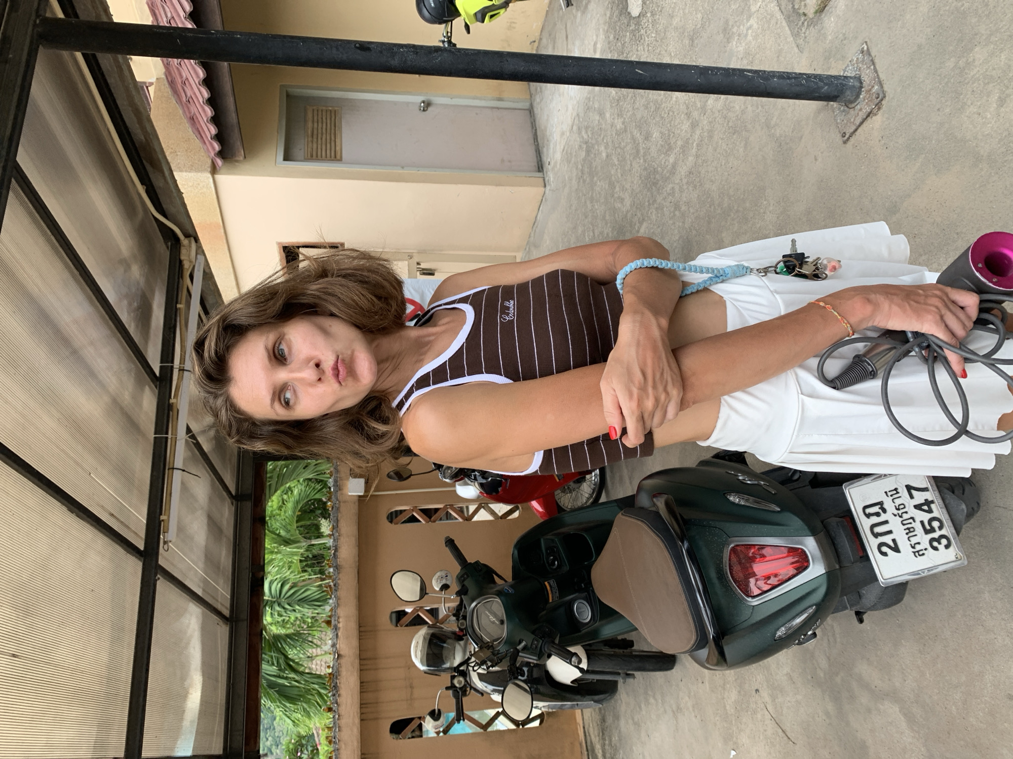 A woman stands in front of a row of scooters outside a building with a covered walkway. She is wearing a brown and white striped tank top and a white skirt. Her hair is brown and wavy, and she is looking off to the side with her lips pursed. She is holding a hairdryer in her right hand and a set of keys on a blue lanyard in her left. The scooters appear to be parked in what looks like a residential or commercial area, with a beige building behind them and palm trees visible in the background. The ground is paved with concrete. The license plate of the closest scooter is visible and appears to have Thai characters. This scene is consistent with an outdoor area in Baan Bo Phut, Thailand, likely a parking area or the entrance to a property.