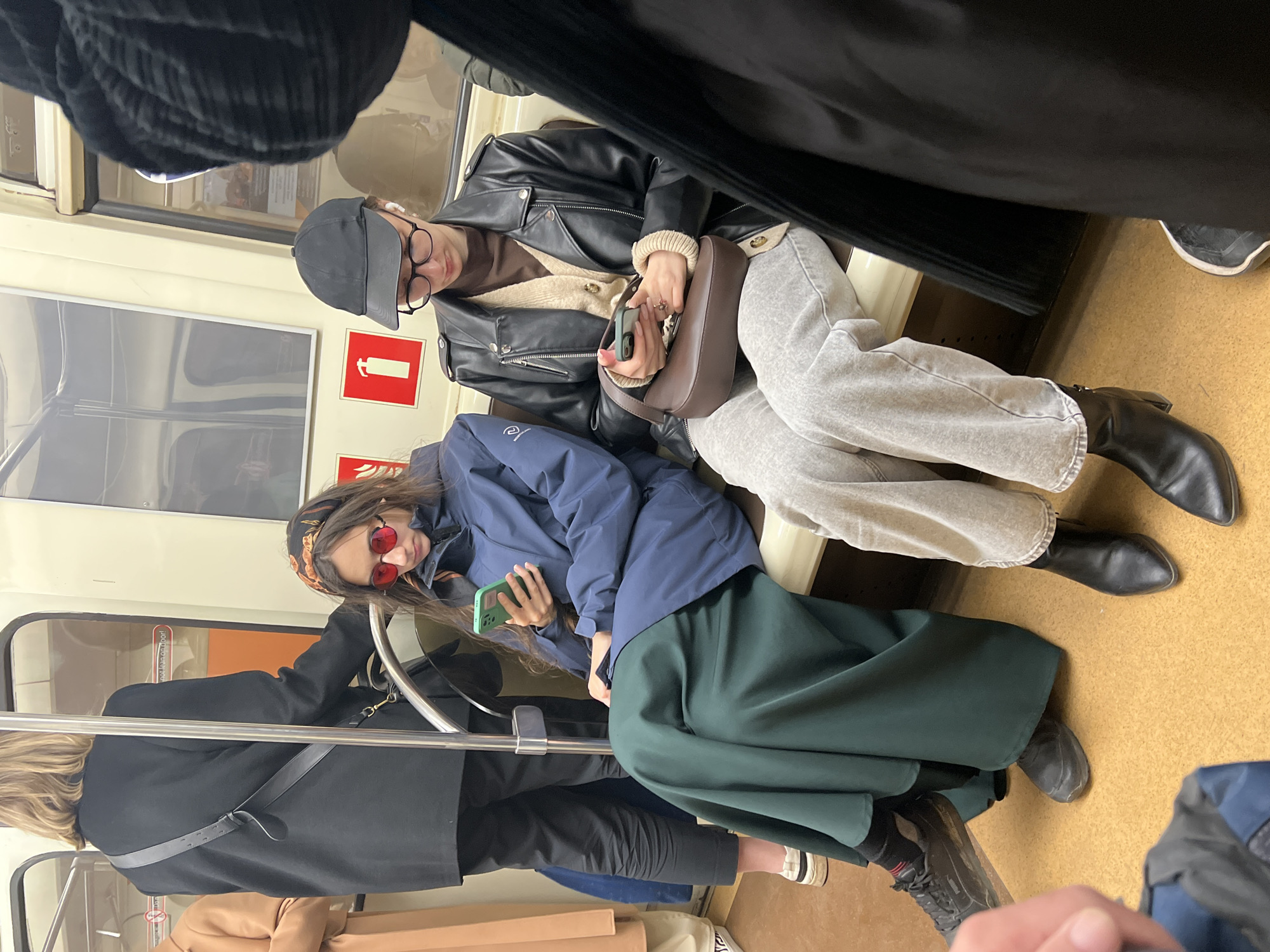 The image depicts the interior of a subway car, likely in Saint Petersburg, Russia, as indicated by the subway car's design elements. Two women are seated on a bench running along the side of the subway car. The woman on the right wears a black leather jacket over a light-colored sweater, gray trousers, and black ankle boots. She has a black cap on her head and glasses, and she's holding a brown purse and a smartphone in her hands. The woman on the left wears a blue jacket over a long green skirt and is holding a smartphone in her hand. She also has red sunglasses and a headscarf. The interior of the subway car is visible, including windows, walls, and handrails. Other passengers are partially visible, standing or sitting in the background. The overall atmosphere suggests a typical urban transportation scene.