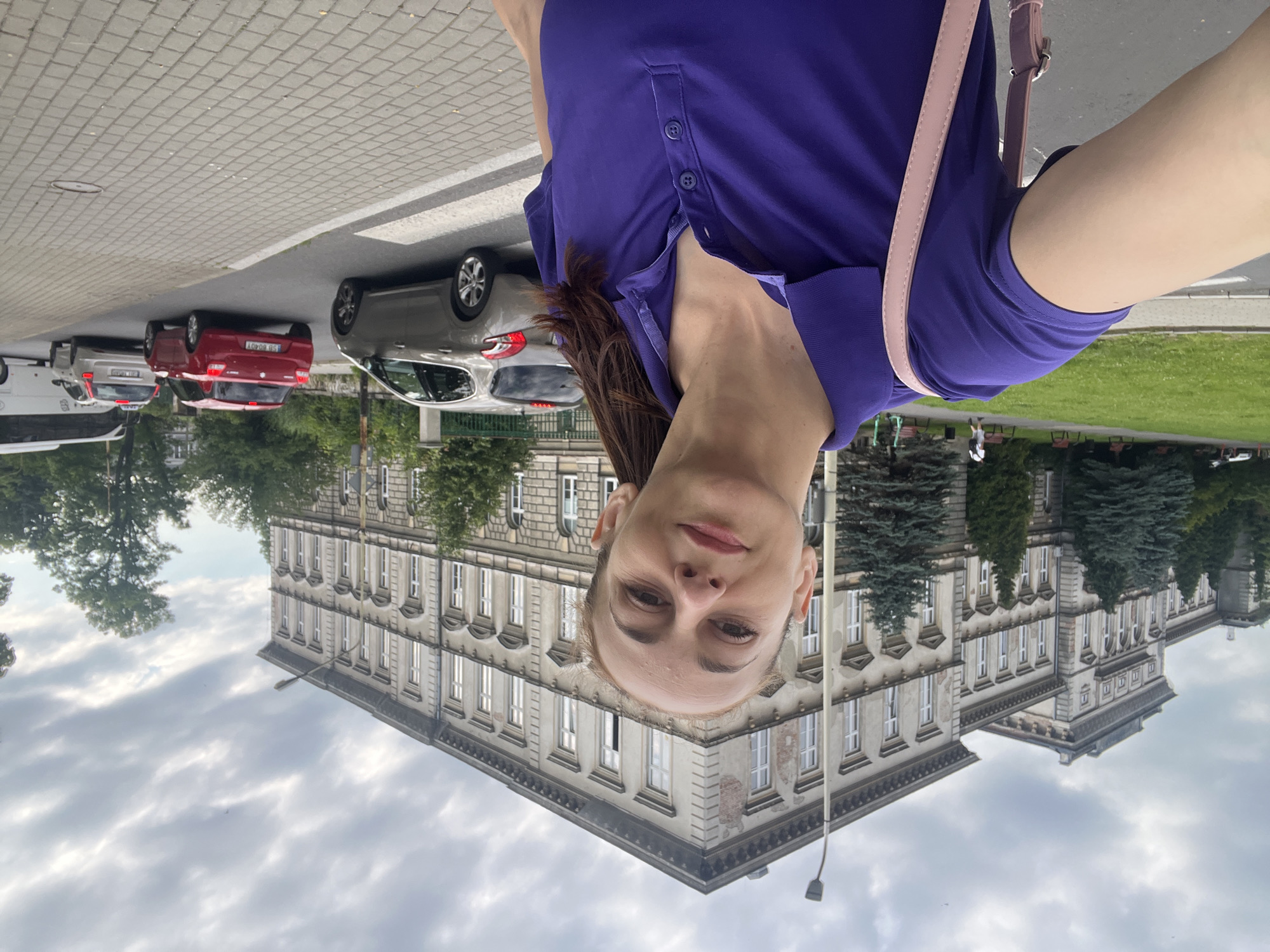 In a selfie taken outdoors on a cloudy day in front of a grand, old building with multiple floors and many windows, a woman with brown hair pulled back is smiling. She is wearing a purple polo shirt and a pink strap from a bag is visible over her shoulder. The building appears to be a school or administrative building with ornate architectural details. In the background, a street is visible with several cars, including a red hatchback, a silver hatchback, and a white bus. The sidewalk is paved with gray bricks. Trees and a grassy area are also present in the scene.