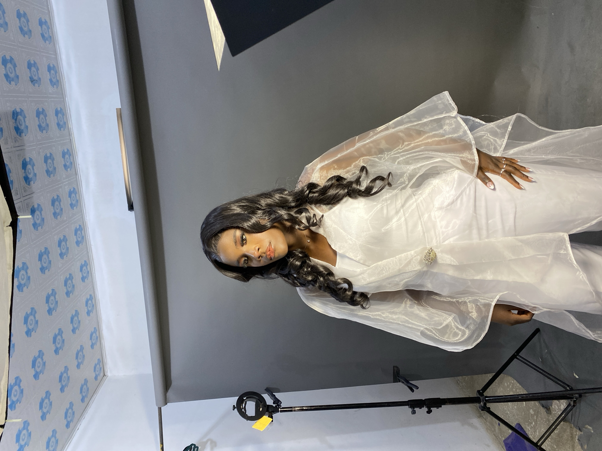 A woman with long, dark, curly hair, is wearing a white gown with a sheer, flowing overlay. She has dark skin, is wearing rings, and is in what appears to be a photo studio, given the lighting equipment. The background consists of grey and white walls, along with blue patterned tiling. The presence of light stands and a dark grey backdrop indicates a controlled studio environment. There's no explicit information to confirm if the location is Jos, Nigeria.