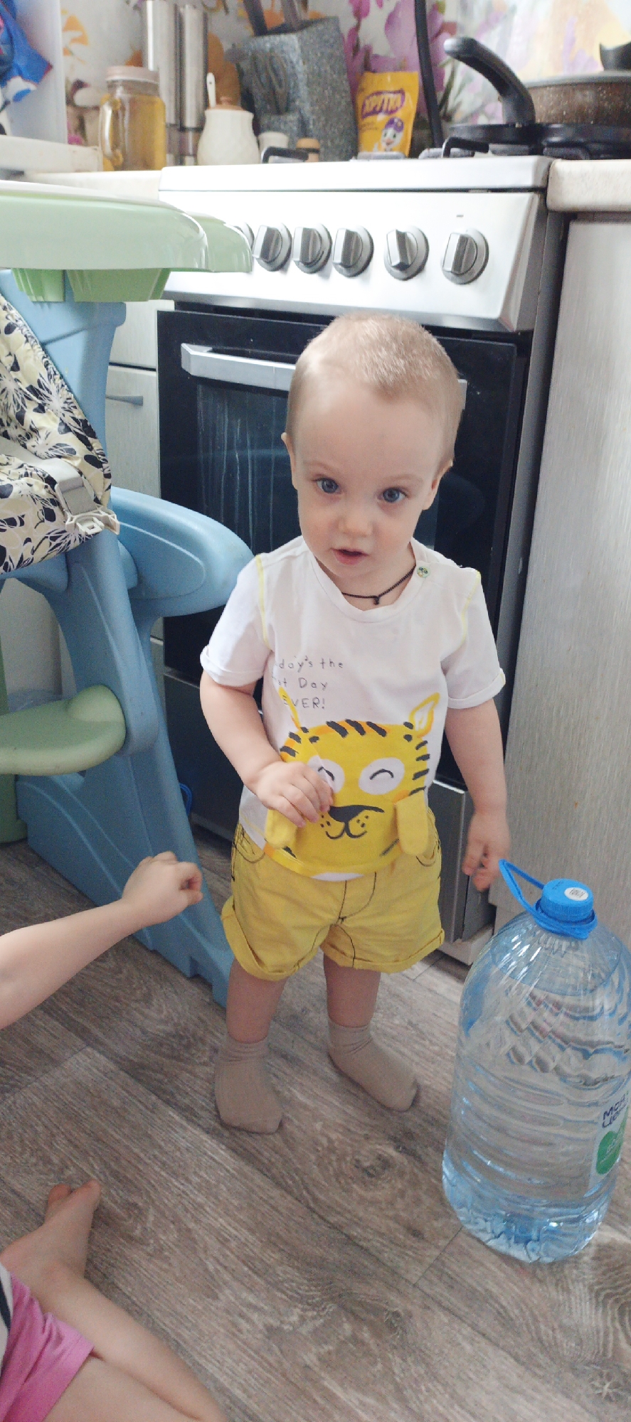 A young child stands in a kitchen setting. The child is fair-skinned, with a light blonde buzz cut and blue eyes, wearing a white t-shirt featuring a cartoon tiger and yellow shorts with khaki socks. The child is standing near a large bottle of water with a blue cap and handle. To the left is part of a child’s blue high chair. An arm and foot of another child are visible in the lower left corner, with the visible foot wearing a pink garment. Behind the child is a white gas range. Above the range are various kitchen items. The floor appears to be wood-grained laminate. The location is identified as being in Chaikovsky, Russia.