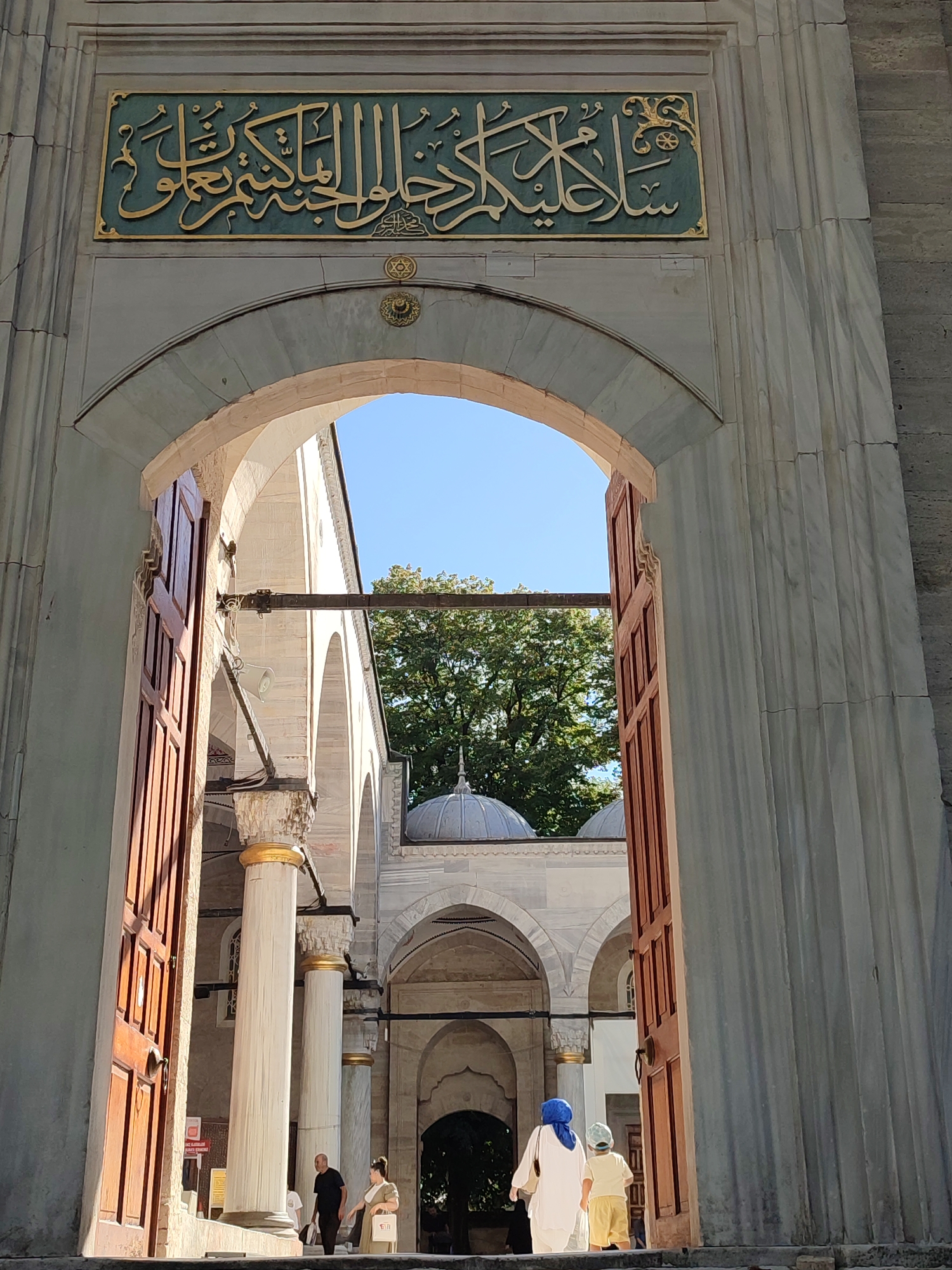 The image shows the entrance to the Rüstem Pasha Mosque, a historic Ottoman mosque located in the Üsküdar district of Istanbul, Turkey. The grand arched entrance is flanked by tall, fluted marble columns. Above the arch, a plaque displays intricate gold Arabic calligraphy on a teal background, a common decorative element in Ottoman architecture. Sunlight illuminates the scene, casting shadows and highlighting the textures of the stone and wood.
Through the archway, one can see a courtyard with more arches and domes, characteristic of mosque architecture. Lush green trees are visible in the background, suggesting a pleasant outdoor setting.
In the foreground, a few people are seen walking through the entrance. One person, possibly a woman, is wearing a blue headscarf and a white robe. Another individual, a child, is wearing a light-colored t-shirt and yellow shorts. They appear to be entering or exiting the mosque complex. The wooden double doors of the entrance are partially open, revealing more of the interior courtyard. The overall impression is one of historical grandeur and peaceful religious ambiance.