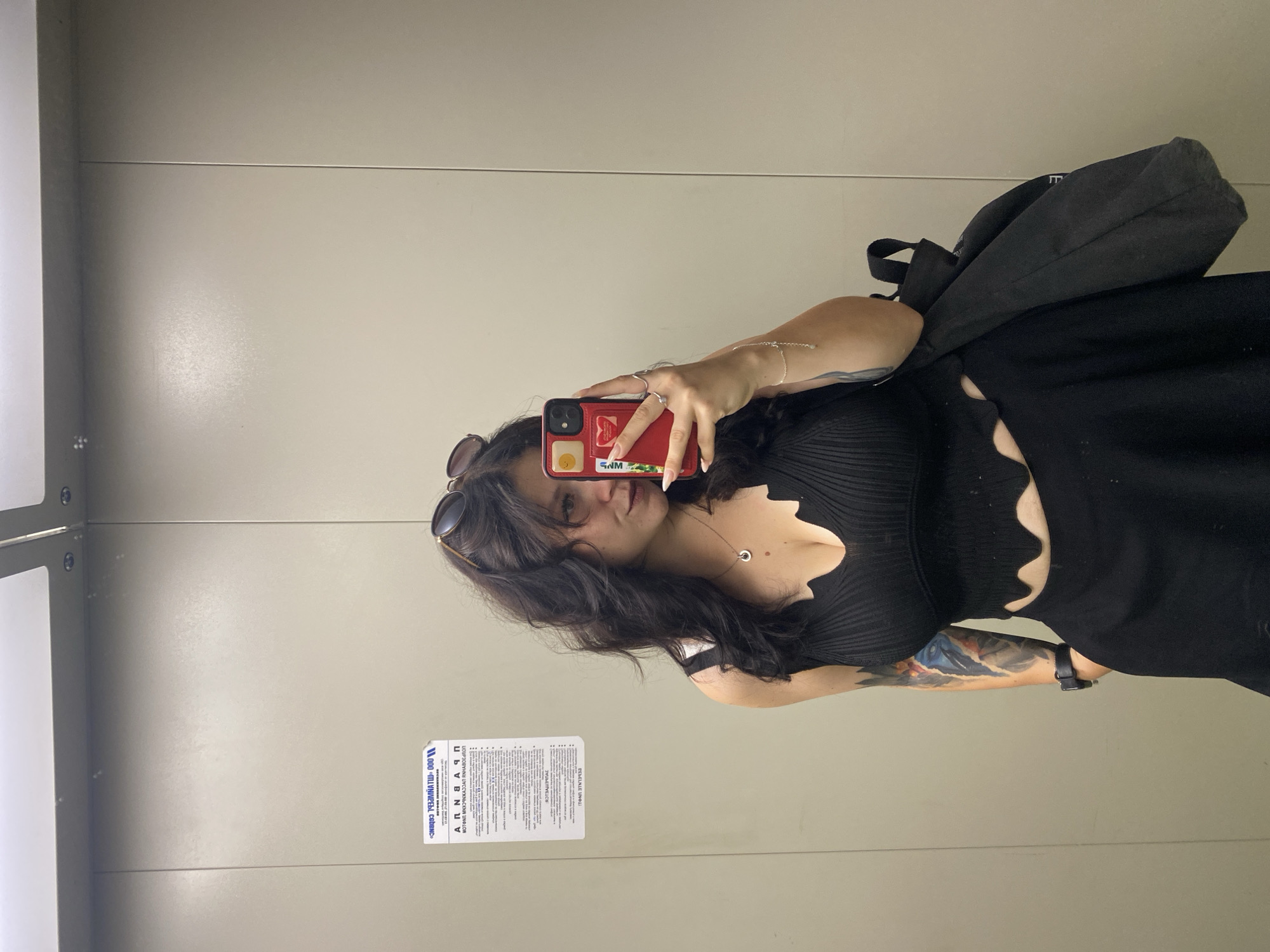 A young woman with dark, wavy hair and tattoos on her arm is taking a selfie in an elevator. She is wearing a black ribbed crop top and a black skirt. She has sunglasses perched on her head and is holding a red smartphone in her right hand. A black backpack is slung over her left shoulder. The elevator walls are a light beige color, and there is a blue and white sticker with text in Russian on the left wall. The scene appears to be in an elevator in Yanino-1, Russia.