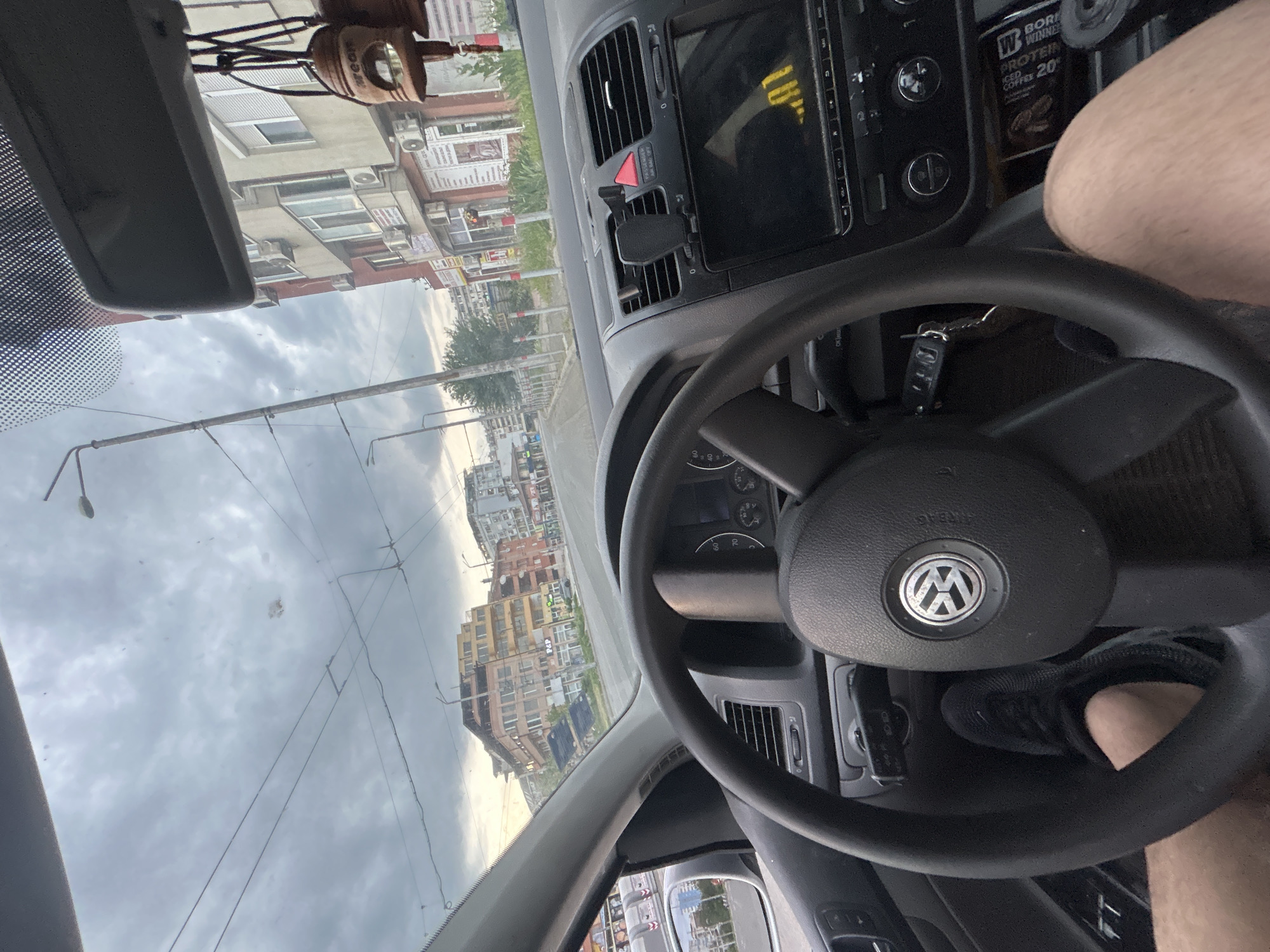 The image depicts a view from inside a car, likely a Volkswagen, in Burgas, Bulgaria. The car is facing down a street lined with buildings, which are visible through the windshield. The sky is overcast. Inside the car, the steering wheel is prominently featured with the Volkswagen logo visible. The car keys are visible on the steering column. A can of "Born Winner" iced coffee is in the front of the car and the driver's leg is visible behind the wheel and gearstick. The driver is wearing black shoes.