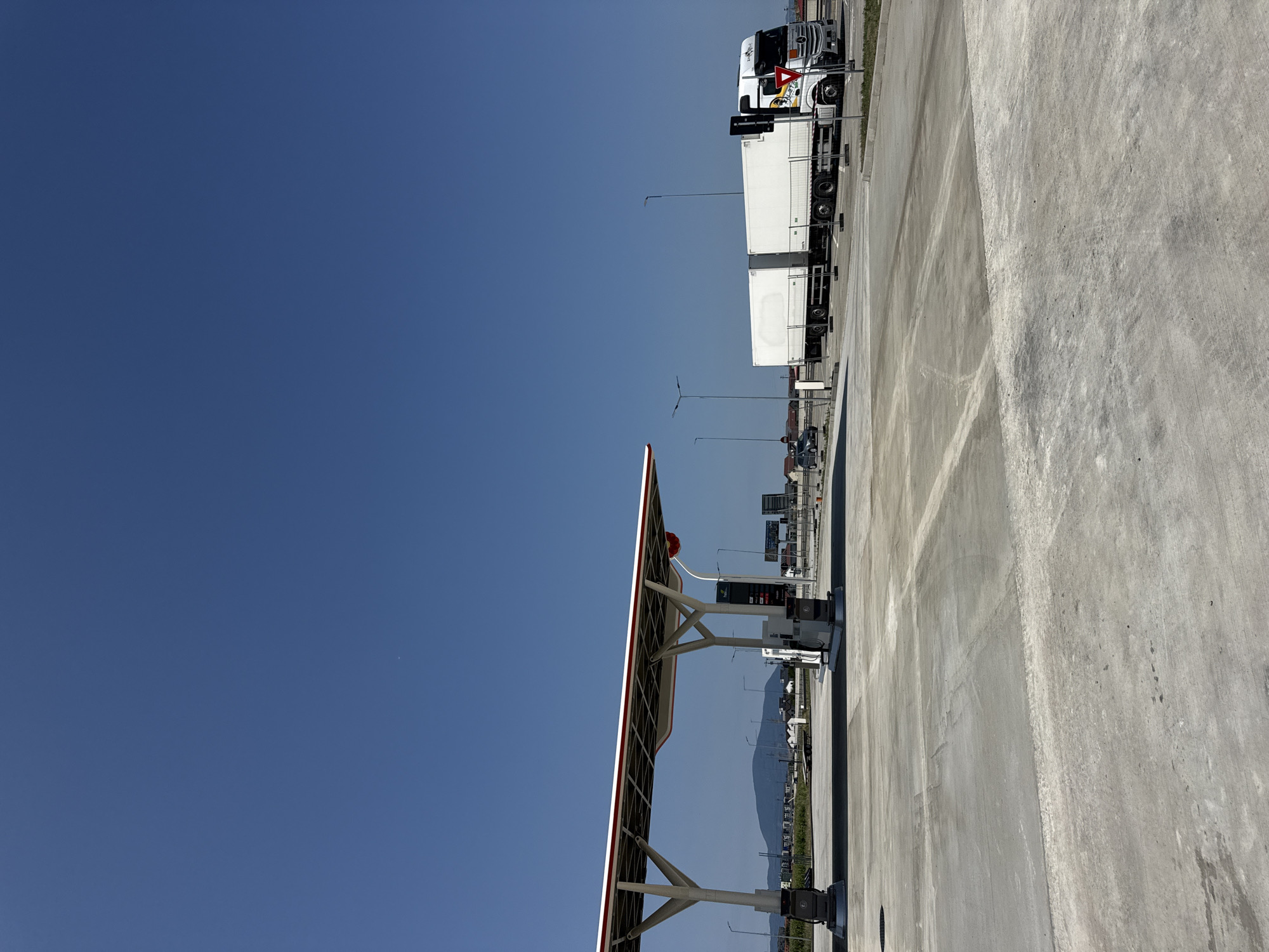 The image depicts a sunny day at a modern gas station in Sanpetru, Romania. The scene is dominated by a clear blue sky and a vast expanse of concrete ground. In the foreground, the concrete surface shows some tire marks and minor stains. A covered gas station canopy with white supports and a red trim is visible on the left, with gas pumps beneath it. In the middle ground, a white semi-trailer truck is parked on the right, its trailer extended. The truck has some logos and text on its side, including what appears to be "SIL-STAR" in black lettering. A small, dark-colored car is parked on the left side of the road in the middle ground. A few streetlights are dotted around the area. In the background, a low, green hill can be seen, with some buildings and vegetation in between. The overall impression is of a functional and well-maintained service area.
