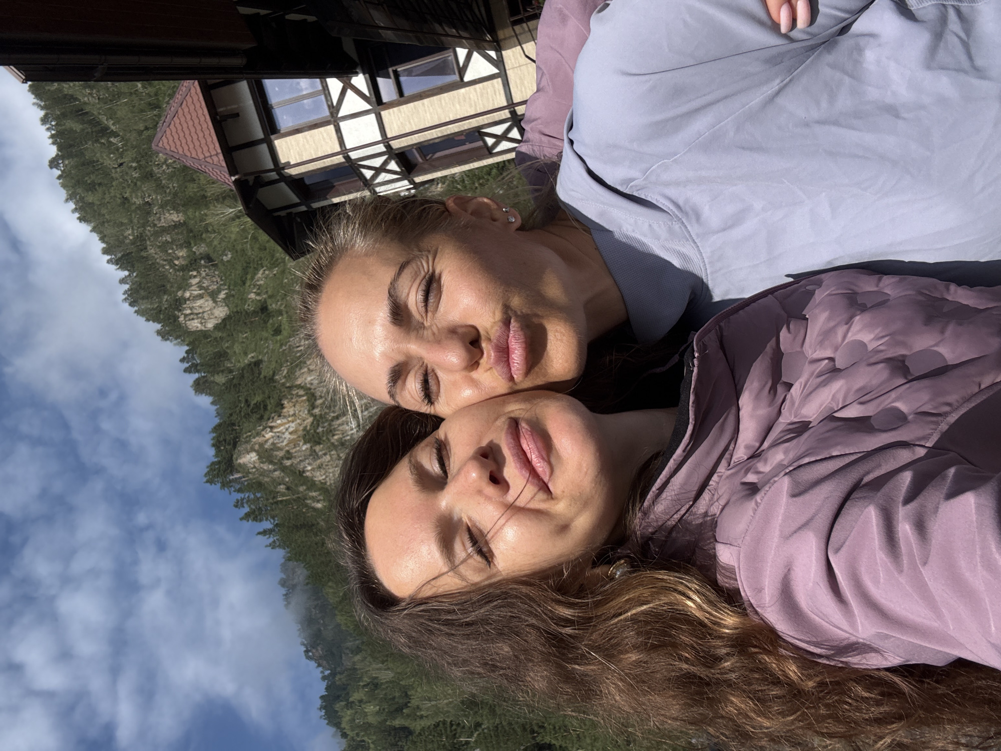 Two women are taking a selfie outdoors in front of a building and a mountain. The woman on the left has long, dark, wavy hair and is wearing a purple puffer jacket. She is smiling with her eyes closed and her mouth slightly open. The woman on the right has blonde hair pulled back and is wearing a light blue hooded jacket. She is puckering her lips for a kiss and has her eyes closed. The background shows a traditional alpine-style building with a red tiled roof, a mountain covered in trees, and a blue sky with clouds. This scene is located in the city of Barangol, Russia.