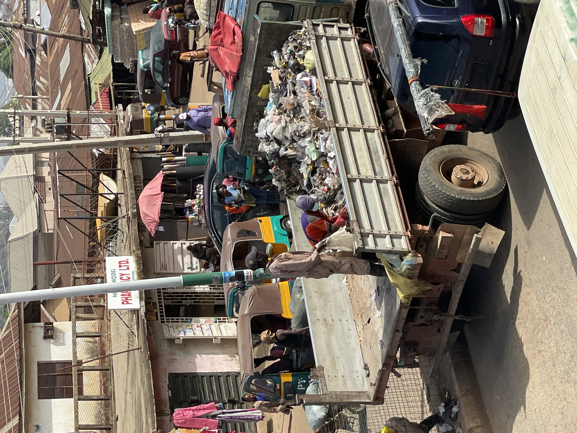 In Bukuru, Nigeria, a garbage truck with a full load of mixed waste is parked on a street. The truck's contents include plastic bags, paper, and other debris. Several people are near the truck; some appear to be sorting through the garbage. A blue car is parked next to the garbage truck, and a spare tire sits on top of the truck's chassis. Behind the truck and across the street are buildings of various sizes and materials, including corrugated metal and concrete. A sign for "New Model Pharmacy LTD" is visible on one of the buildings. Tuk-tuks are parked along the street.