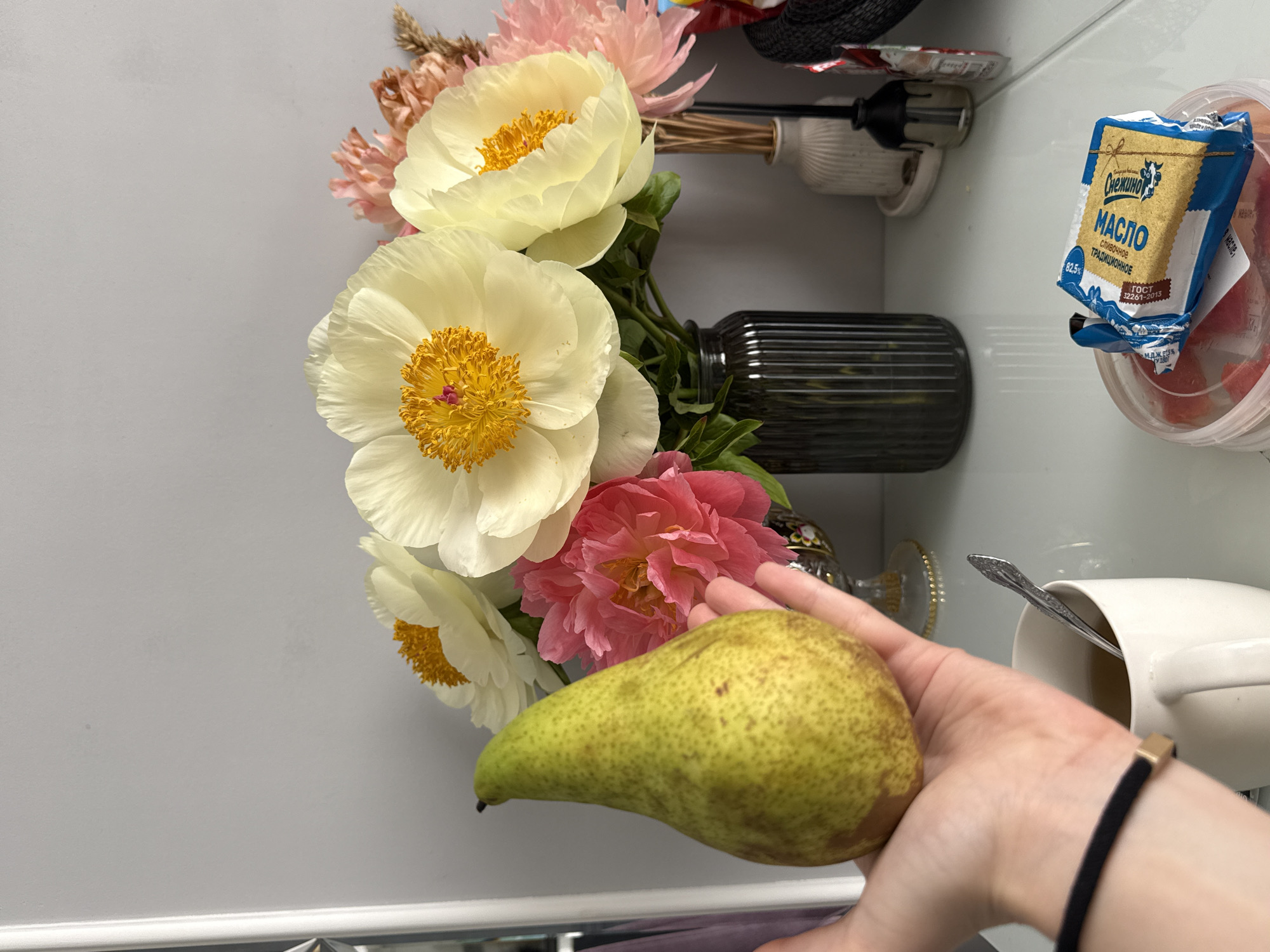 A photograph depicts a still life arrangement set against a white, possibly reflective, surface. A bouquet of light yellow and pink peonies is arranged in a dark glass vase with horizontal ridges. To the right of the vase is a partially visible package of butter labeled with Cyrillic text, positioned on top of a round, clear container. A white mug with a spoon inside rests further to the right. In the foreground, a hand holds a green and speckled pear. The environment is an interior space, likely a kitchen or similar area.