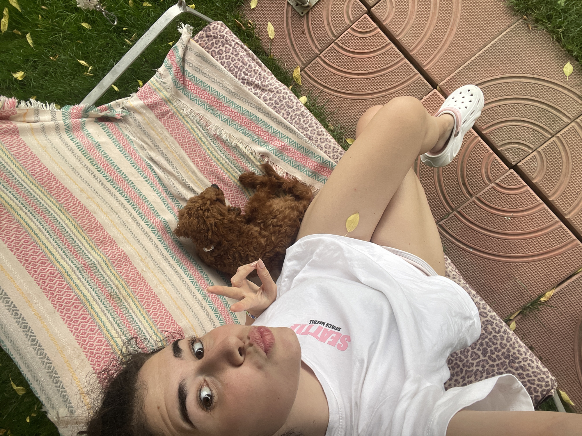 A young woman is lying on a portable cot with a brown poodle nestled next to her on a striped blanket. She is wearing a white t-shirt and a white shoe on her right foot. A yellow leaf rests on her leg. The cot is situated on a mix of green grass and red-brown tiled surfaces, potentially a patio. The image might be from Sofia, Bulgaria, but without additional context, pinpointing the exact location is speculative.