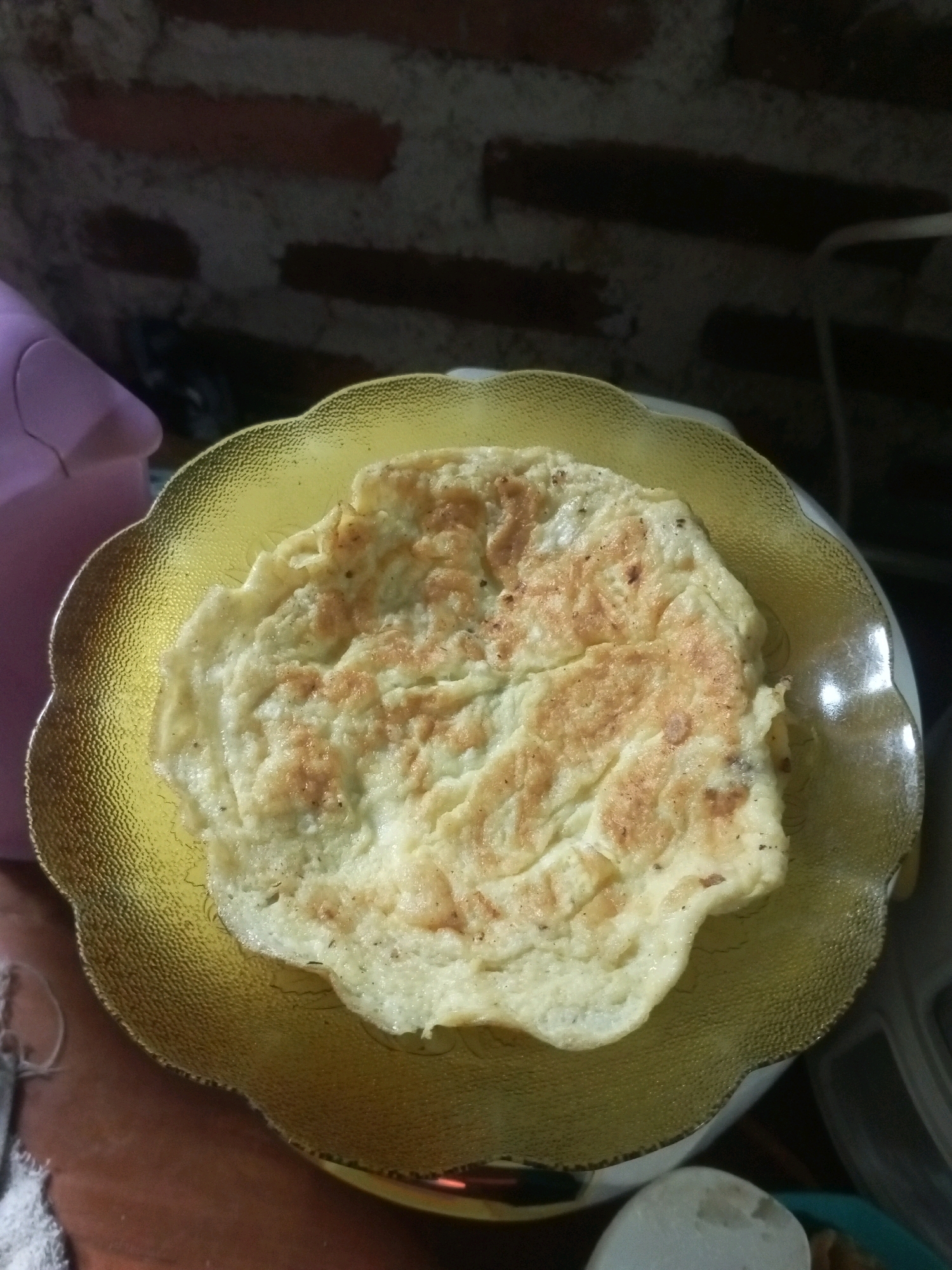 A close-up, overhead shot shows a cooked egg omelet on a decorative gold-colored plate. The omelet is unevenly browned with darker cooked edges and lighter, fluffy centers. The plate has a scalloped edge and a textured surface. In the background, a brick wall with visible mortar lines and a pink object, possibly a kitchen appliance, are dimly visible. The overall lighting is dim, suggesting an indoor setting. The scene appears to be in a domestic environment, possibly a kitchen, in Cimarga, Indonesia.