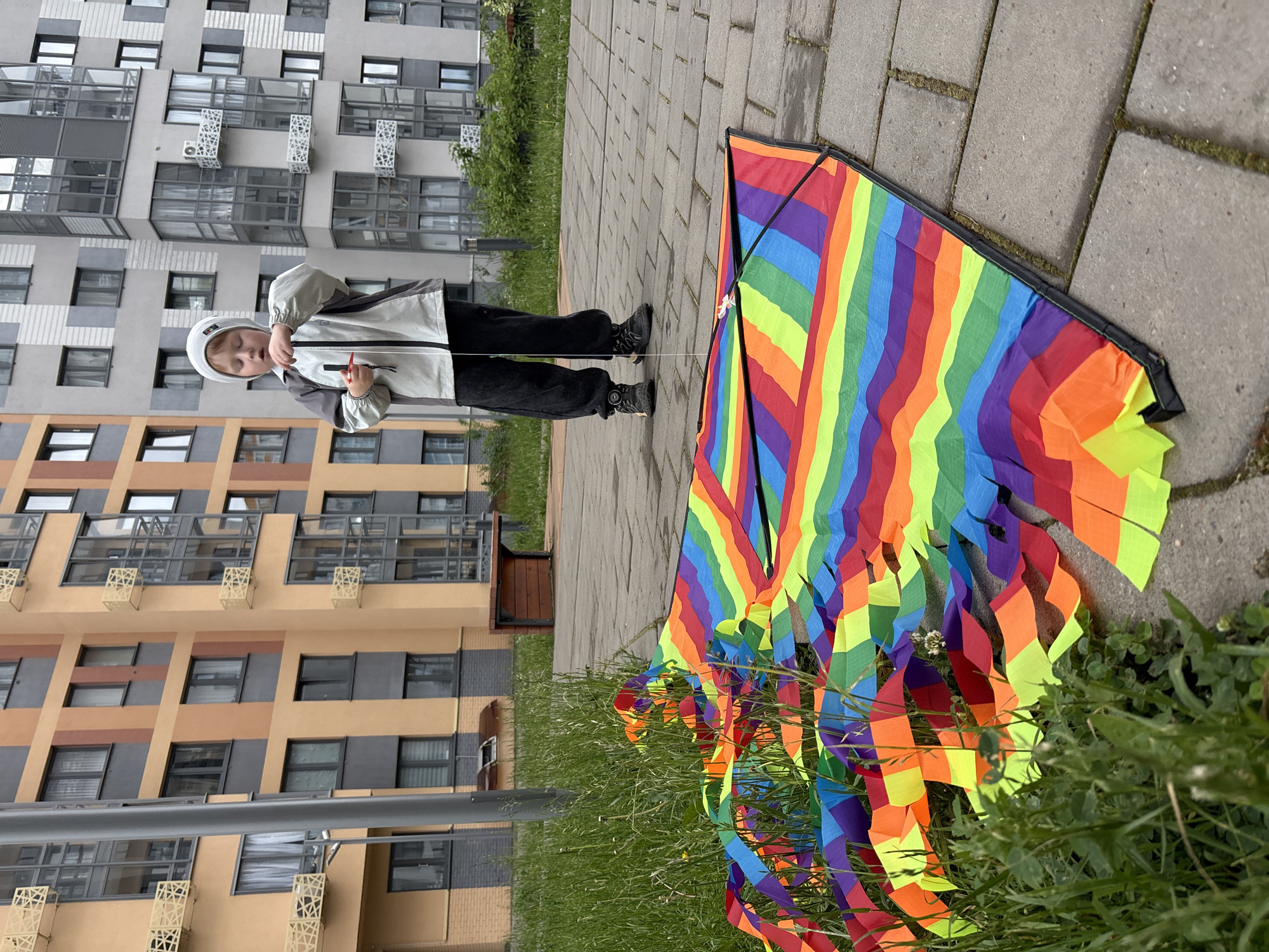 The image depicts a young child, approximately 4-7 years old, standing on a grey paved walkway next to a large, multi-colored kite in a residential area of Izhevsk, Russia. The child wears a two-tone jacket with a light grey upper and darker grey lower section, black pants, and dark sneakers, topped with a light grey or white beanie. They are holding a black and red kite string winder, around which white string is wrapped, with both hands at chest level, and their gaze is directed downwards, focused on the winder or the kite.
The kite, a delta-style design, is laid flat on the paved surface, with its long, streamer-like tail extending into an adjacent strip of tall green grass. The kite features prominent horizontal stripes in the colors of the rainbow: red, orange, yellow, green, blue, and purple. Its tail is composed of multiple vertical strips of the same vibrant colors, creating a ruffled appearance. Black framing rods are visible on the kite's structure. The paved walkway consists of rectangular grey stones, with some moss or dirt present in the crevices.
In the background, modern, multi-story residential buildings dominate the scene. The building on the left is characterized by light orange/peach and dark grey paneling, featuring rectangular windows and decorative, geometric-patterned metal grates or railings. The building to the right and center background is primarily light grey with dark grey sections, exhibiting similar contemporary architectural elements, including glass-enclosed balconies or bay windows. A dark grey cylindrical pole, possibly a street lamp or utility pole, stands on the left side of the frame, partially obscured by foreground grass. A wooden bench with a dark metal frame is positioned in the mid-ground, near the base of the buildings. The scene is illuminated by soft, even light, indicating an overcast day, and the lush green grass suggests late spring or summer.
