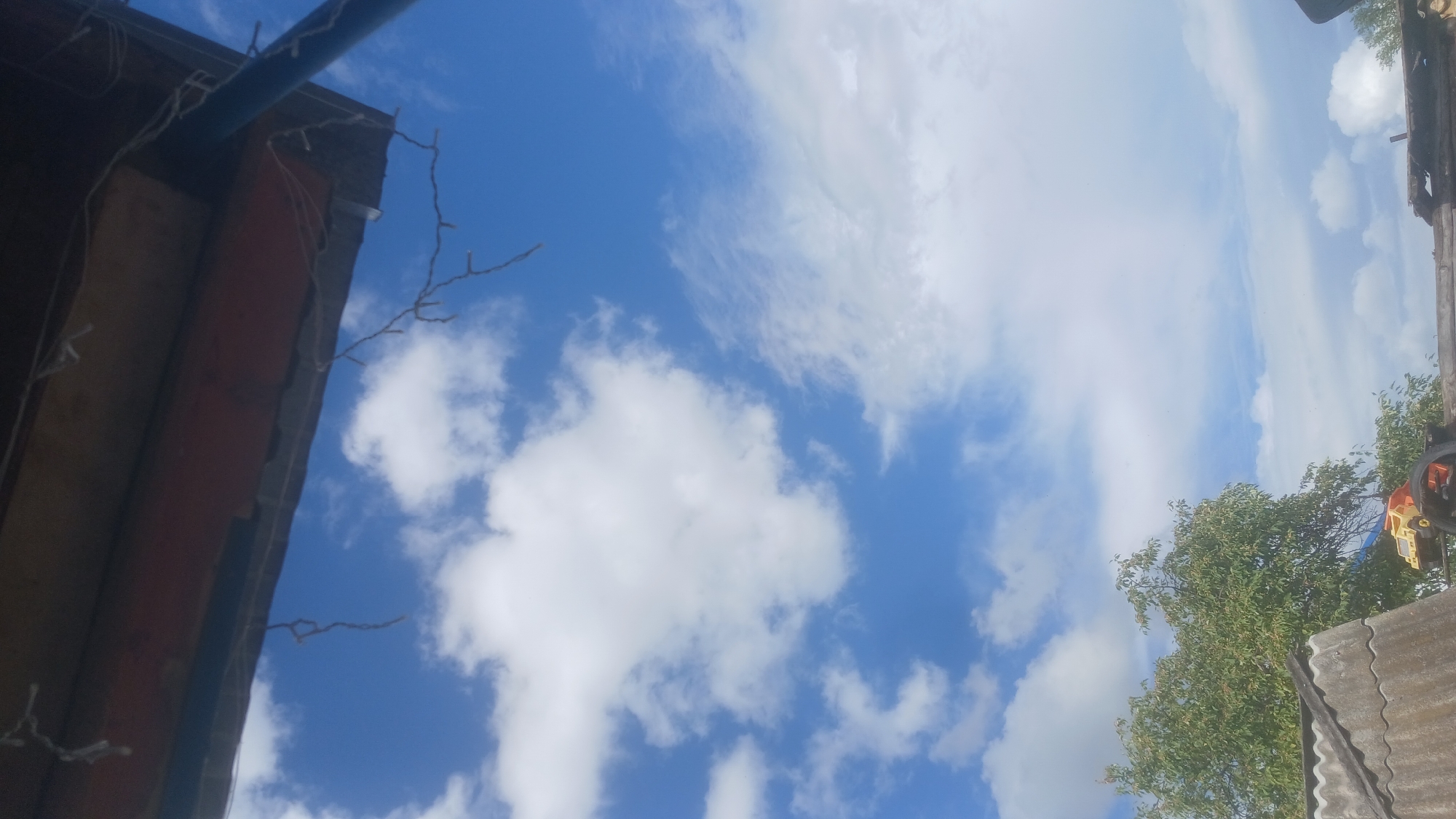 The image depicts a cloudy blue sky with a view looking upwards, suggesting it was taken from a low vantage point. In the foreground, part of a wooden structure with some hanging string lights is visible, possibly an overhang or a roof. Below this, to the left, is a gray, corrugated roof. To the right of the corrugated roof, a vibrant green tree with dense foliage is present, with a small yellow and orange toy truck visible amongst some debris, possibly on a tire, in front of the tree. The overall scene suggests an outdoor environment, possibly a backyard or a less developed area. The image does not provide enough information to determine the specific location within Slak, Russia, or to identify any individuals or specific actions taking place.