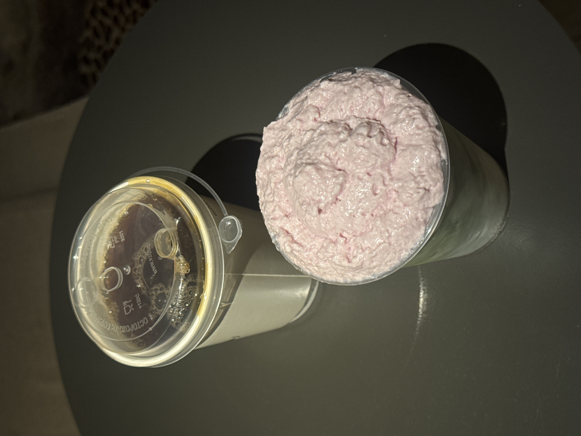 Two plastic cups sit on a dark, circular surface, likely a table. The cup in the foreground is filled with a light pink, frothy substance that mounds over the rim. The cup behind it is partially obscured by the first cup and contains a dark liquid topped with a clear plastic lid. The lid is slightly ajar, revealing some condensation and bubbles in the liquid. The surface the cups are on is smooth and reflects some light, casting shadows from the cups. The background is dark and out of focus, with subtle textures suggesting a dimly lit environment.