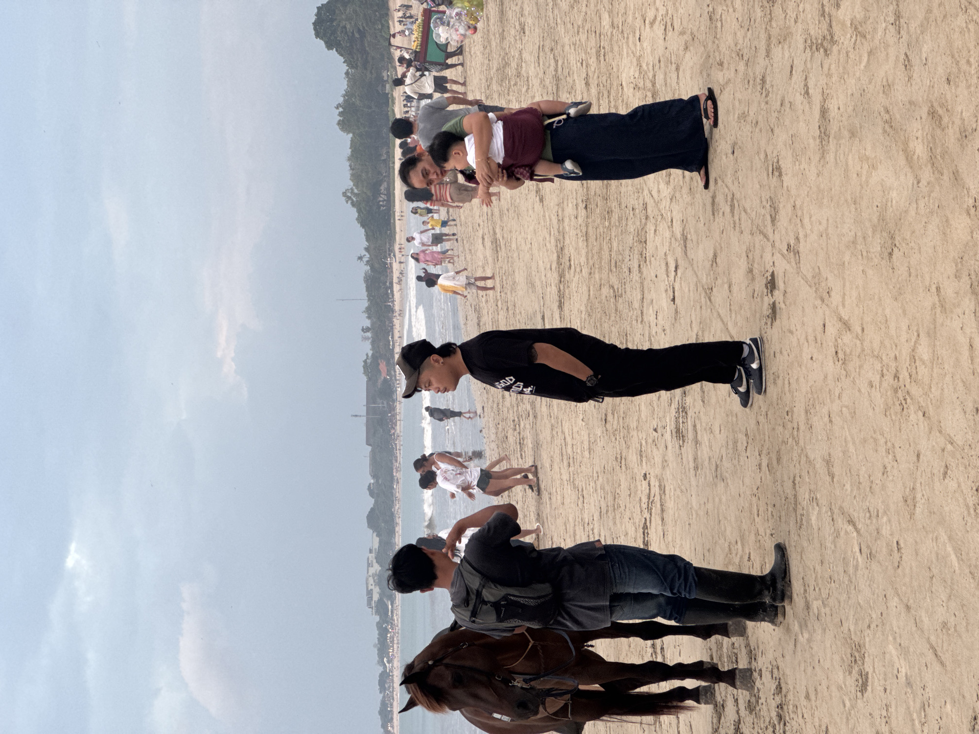 The image shows a beach scene in Kuta Selatan, Indonesia, with several people and a horse. The sky is overcast, suggesting a cloudy or hazy day. The sand is light brown and appears to be damp in some areas, indicating proximity to the ocean.
In the foreground, a person is riding a brown horse. The rider is wearing a dark grey t-shirt, blue jeans, and black boots. The horse's mane is light brown.
Further up the beach, a person in a black t-shirt and black cap is standing. Behind them, a group of people are walking and standing. One person is holding a young child. Several other individuals are scattered across the sand, some appearing to be walking towards or away from the water. In the distance, the ocean waves are visible, and a line of trees borders the far side of the beach.
There are no explicit indications of the time of day, but the overcast sky suggests it is daytime. The overall mood appears casual and relaxed, typical of a beach environment. No visible text is discernible in the image.