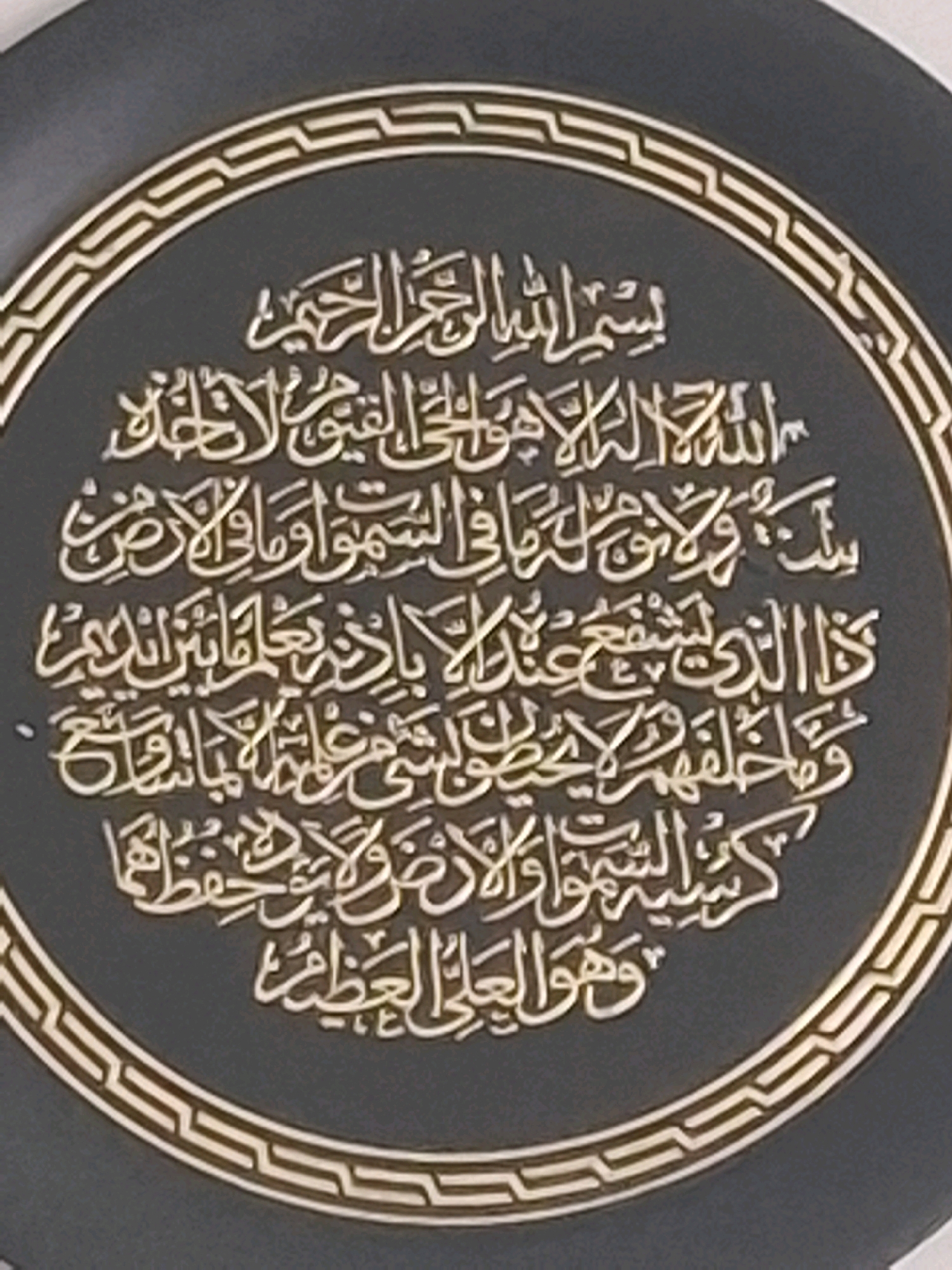 The image shows a decorative plate with Arabic calligraphy. The calligraphy is golden and set against a dark grey background. The plate is surrounded by a golden border with a geometric pattern. The scene is a close-up, with no environmental context provided. It is not possible to determine the location in the city of Almaty, Kazakhstan from this image.