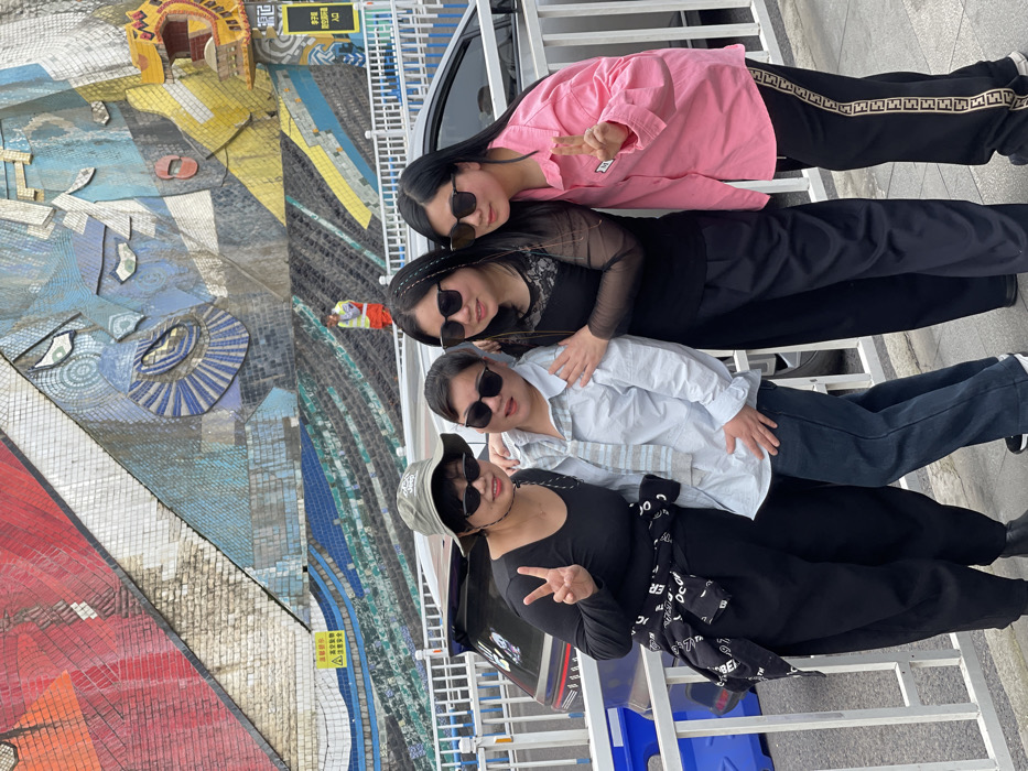 The image depicts four adult females posing for a photograph on a paved sidewalk in Yuzhong District, China. From left to right: the first individual wears a khaki bucket hat, black cat-eye sunglasses, a black long-sleeved top, black baggy pants with a patterned shirt tied at the waist, and displays a peace sign with her left hand. Her right arm is around the second individual, who wears dark sunglasses, a light blue button-up shirt with a striped element, dark jeans, and has her right arm around the third person. The third individual wears dark square-framed sunglasses, a black sheer-sleeved top, black wide-leg pants, and embraces both adjacent individuals. The fourth individual wears dark square-framed sunglasses, a pink long-sleeved collared shirt, black track pants with a patterned side stripe, and displays a peace sign with her right hand. All four are smiling at the camera.
They stand alongside a white metal barrier. Beyond the barrier, a dark-colored SUV is visible on a road. The background is dominated by a large, multi-colored mosaic mural on a sloped wall, featuring abstract facial elements and geometric patterns in blue, red, yellow, and white tiles. A person in an orange high-visibility vest is visible in the middle ground near the mural. Chinese signage, including a yellow warning sign and a white "入口" (Entrance) sign, is present in the background. The scene depicts an urban setting.