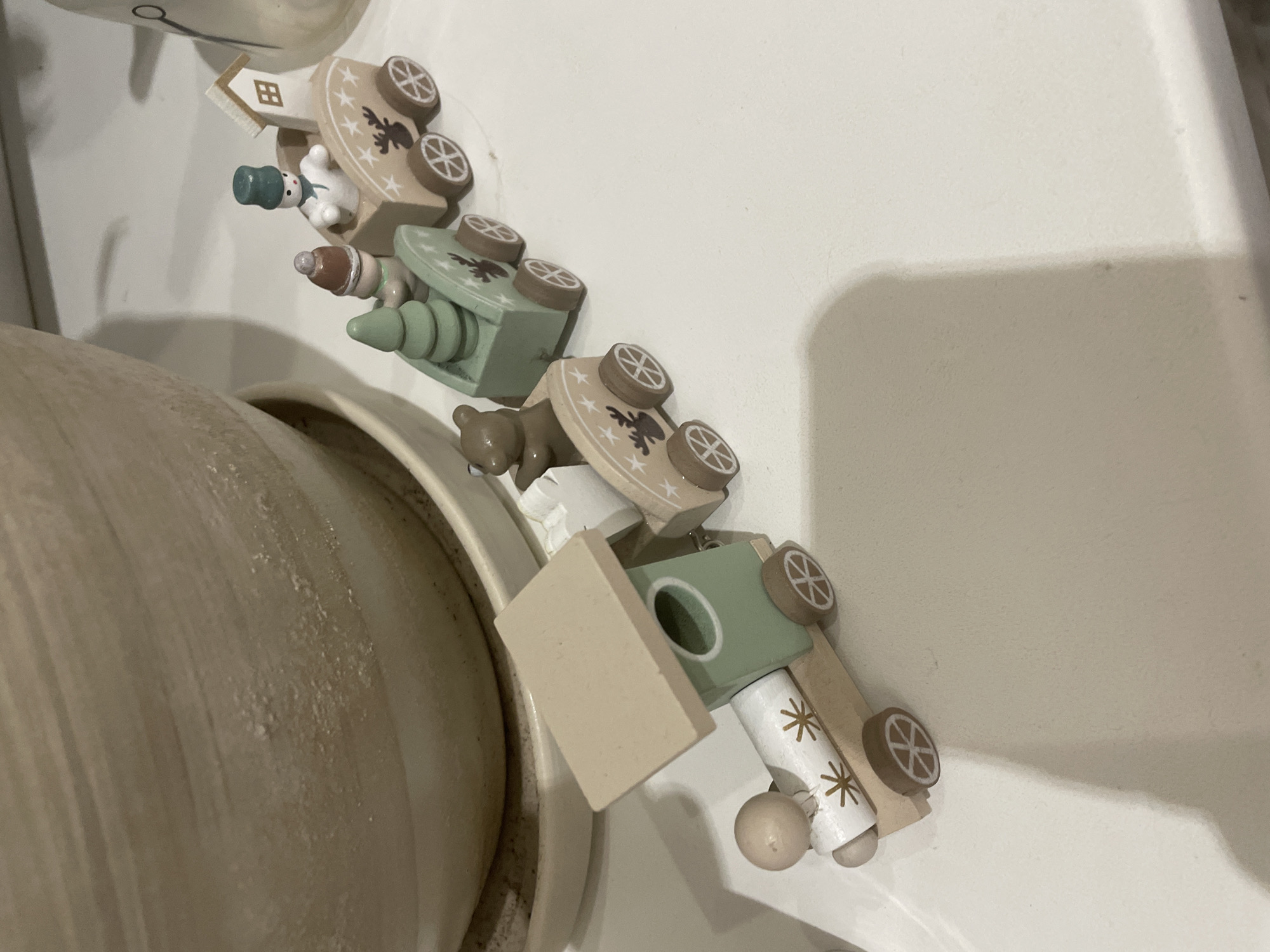 A close-up, elevated shot shows a decorative wooden toy train on a white surface. The train consists of a locomotive and three carriages, each with two wheels. The locomotive is green and tan with a tan roof and has a round, light-brown knob on top. The first carriage is tan with a brown reindeer and star motif, containing a small brown bear figurine. The second carriage is green and has a miniature evergreen tree. The third carriage is tan with a brown reindeer and star motif, and features a small wooden house with a white figure wearing a green hat. Behind the train, a large, textured beige planter is visible. The lighting is dim and diffused, casting a soft shadow from the train onto the white surface. The scene is static and appears to be indoors, possibly a home decoration. The image does not contain any information about the city of Vologda, Russia.