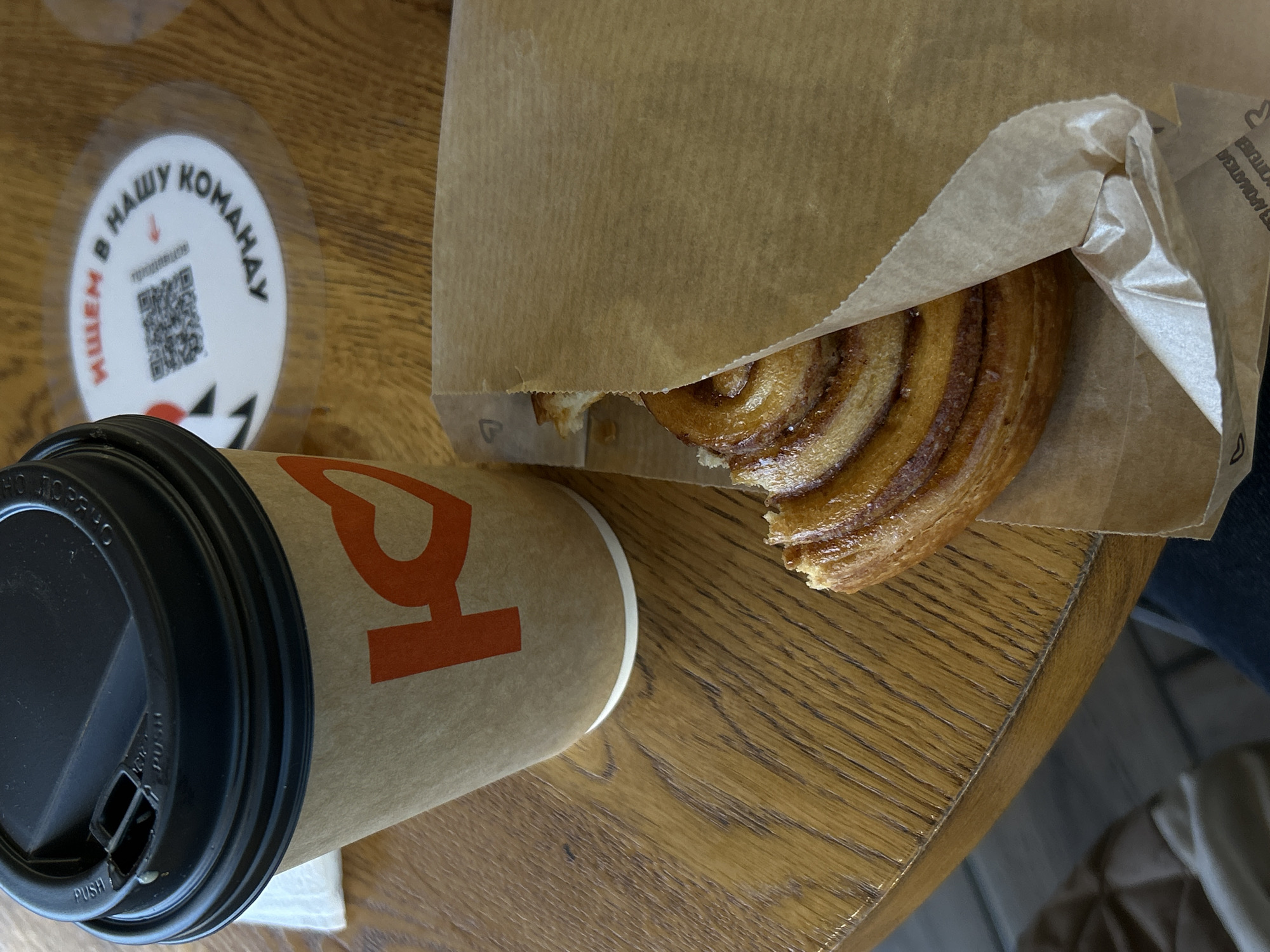 The image presents an overhead view of a coffee and pastry set on a wooden table, likely in a cafe or bakery in Saint Petersburg, Russia. The main subjects are a disposable coffee cup, a pastry in a paper bag, and a sticker on the table.
In the foreground, a light brown paper coffee cup with a black lid is visible. A large, stylized red or orange letter, resembling a Cyrillic "Р" (R) or a heart, is printed on the cup. The black lid features a drinking spout with the word "PUSH" visible near it.
Next to the cup, a glazed cinnamon roll or similar swirled pastry is partially seen, emerging from a light brown paper bag. The pastry appears to be partly eaten, revealing its soft, layered interior. The paper bag is creased, and faint, inverted text is barely discernible on its upper right side, along with a small letter "P" on the bottom left.
On the wooden table, which has a distinct grain pattern, a round, clear sticker is affixed. The sticker features Cyrillic text in black and red, reading "ИЩЕМ В НАШУ КОМАНДУ" (ISHCHEM V NASHU KOMANDU), which translates to "WE ARE LOOKING FOR (PEOPLE) FOR OUR TEAM." Below this text, a QR code is visible, along with a red downward arrow, suggesting a recruitment drive.
The setting implies a casual breakfast or snack, with a person potentially seated at the table, indicated by a glimpse of dark clothing and a light-colored item in the far right background. The image is brightly lit, suggesting daytime. The Cyrillic text on the sticker strongly confirms the Russian-speaking environment consistent with the provided location context of Saint Petersburg.