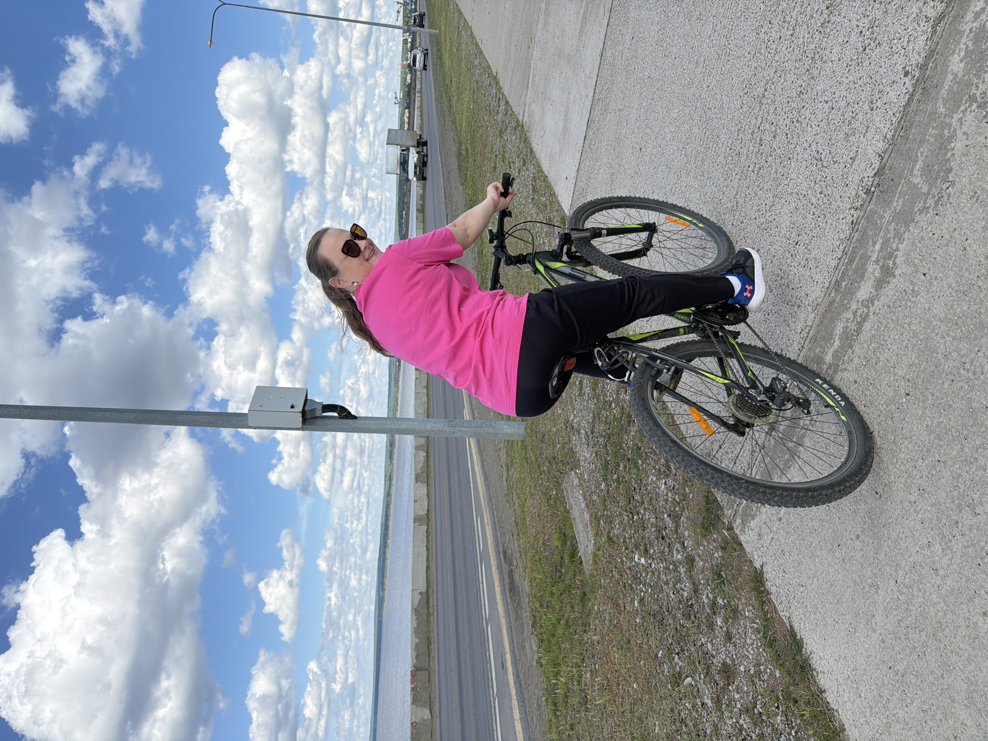 A woman is pictured riding a bicycle on a paved path next to a road. She is wearing a bright pink t-shirt, black pants, and sunglasses. The bicycle has a black frame with green accents and black tires. In the background, there is a body of water, a road with some vehicles, and a cloudy blue sky. A metal pole with a junction box is visible in the foreground. The scene appears to be outdoors during the daytime, under a partly cloudy sky. The woman is looking at the camera and smiling, suggesting a pleasant or happy mood. The text "KENDA" is visible on the tire of the bicycle.