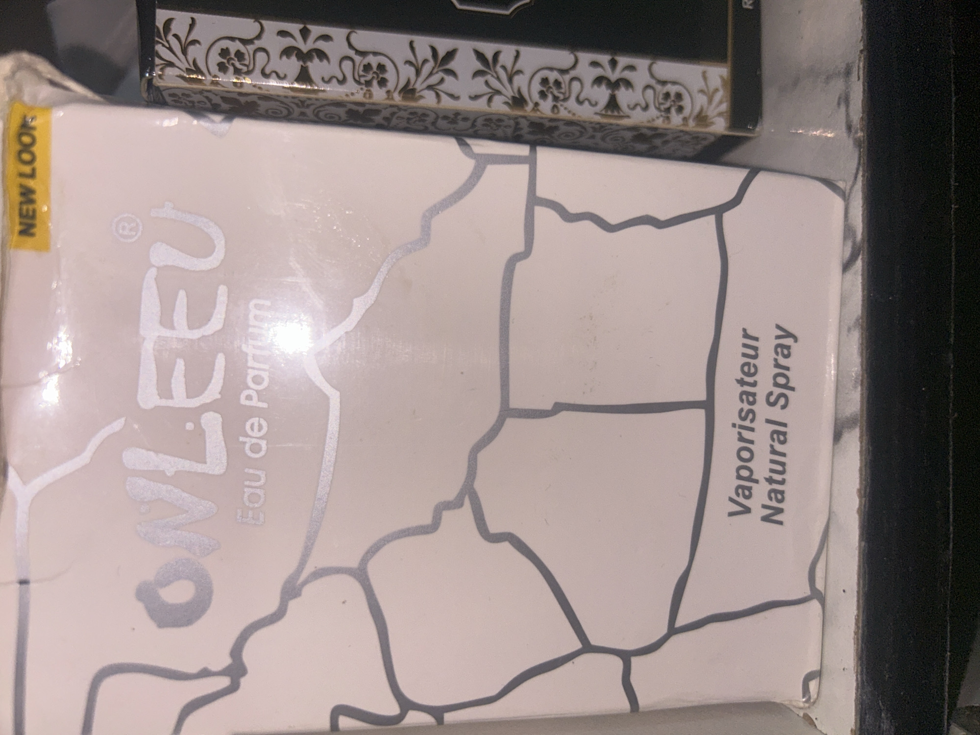 A close-up, eye-level shot shows a product box, likely for perfume, with a cracked pattern design in gray lines on a light cream background. The brand name "ONLEEU" is prominently displayed in a lighter cream font with a subtle sheen. Below the brand name, "Eau de Parfum" is written in a smaller, similar font. In the upper right corner, a yellow sticker reads "NEW LOOK". Towards the bottom of the box, "Vaporisateur Natural Spray" is printed in a darker gray font. The box appears to be sitting on a dark surface, possibly a shelf, with the right side of the image showing part of another product with an ornate black and gold pattern. There are no people or animals visible in the image. The lighting suggests an indoor setting. It is impossible to determine the exact location within Ogbomosho, Nigeria, from the visual information provided.