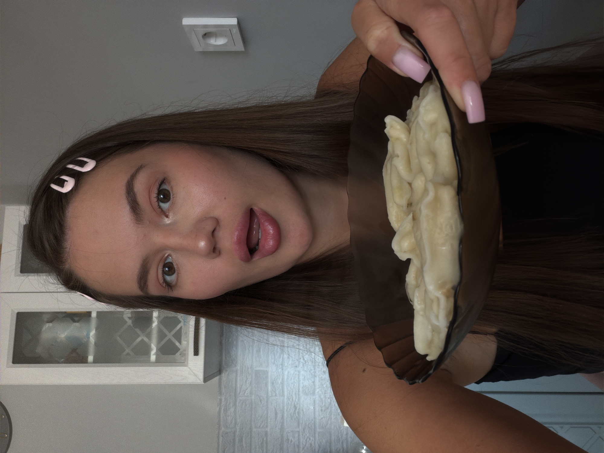 The image shows a young woman in what appears to be a kitchen, with a white brick-patterned wall visible in the background. She is holding a dark brown bowl filled with what looks like dumplings or gnocchi. The woman has long, dark brown hair, some of which falls over her face and shoulders. She is wearing a black top. Two light pink hair clips are visible in her hair. Her right hand, with neatly manicured light pink nails, is holding the bowl. The woman's mouth is open as if she is about to eat or speak, and her expression could be interpreted as playful or surprised. The lighting suggests it might be daytime, but there are no clear indications of the time of day or weather. The presence of kitchen cabinets and a wall outlet indicates an indoor setting. There is no visible text in the image. The location context provided is Sirius, Russia.