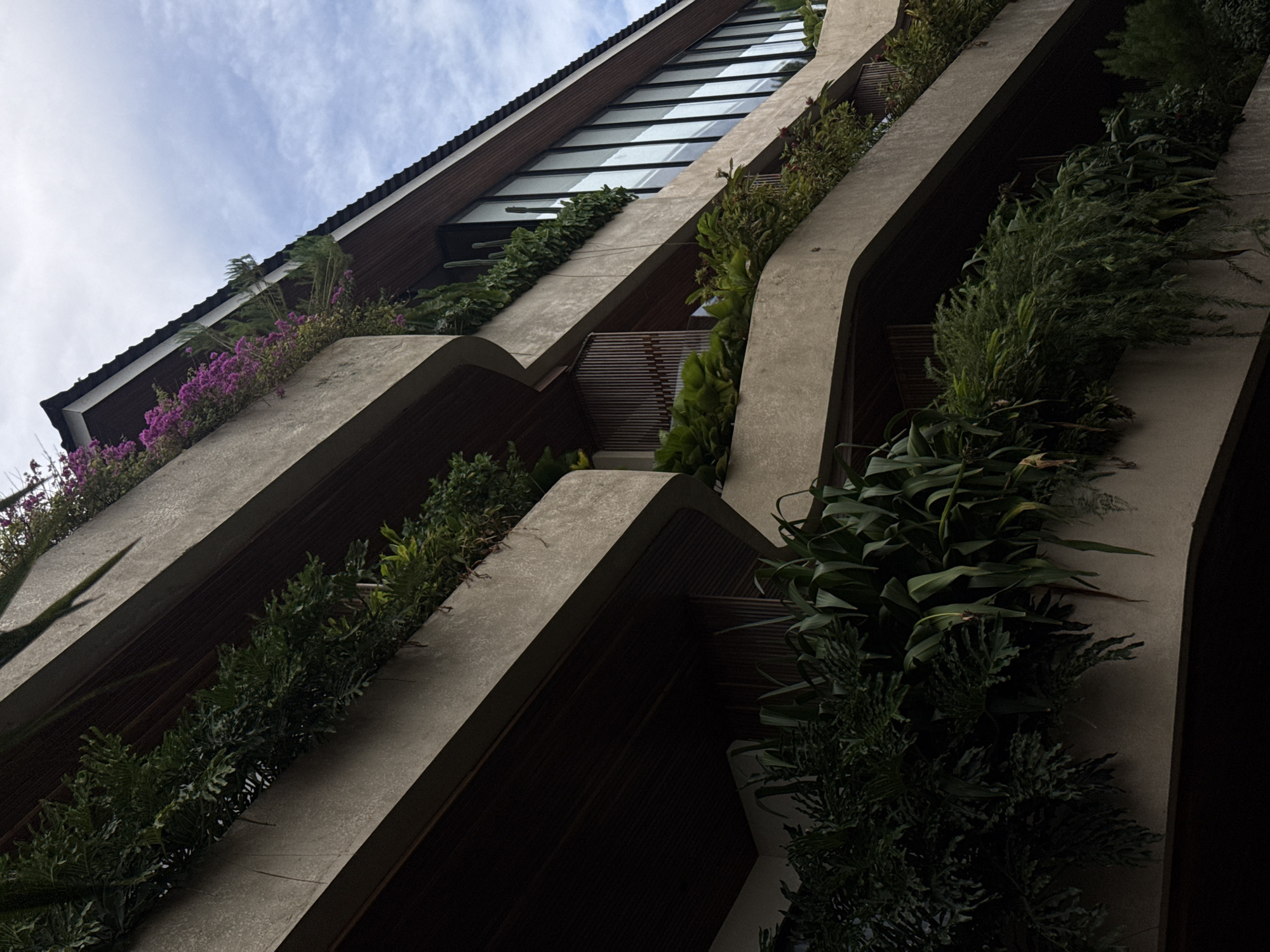 An upward-looking view captures a striking multi-story building featuring biophilic architecture. The structure is characterized by gracefully curved concrete or stone planter boxes that extend horizontally across each level, creating a cascading effect. These planters are densely filled with a variety of lush green vegetation, including ferns, bushy shrubs, and long-leafed plants, contributing to a vibrant vertical garden aesthetic. On the upper levels, bright pink or purple flowering plants add a splash of color against the natural greenery and grey concrete.
The building's facade also includes prominent dark-framed windows, possibly reflective, spanning multiple stories in the upper-middle section, contrasting with the natural elements. A dark tiled roof with a pronounced overhang is visible at the very top, silhouetted against a partly cloudy, light blue sky. Dark wooden slats or panels are visible beneath the planter boxes, providing a warm, natural texture in the shaded areas.
The perspective emphasizes the building's height and the deliberate integration of nature into its design. The varying light and shadow on the concrete surfaces and deep recesses between the levels add depth to the image. The scene is set during the daytime, under a bright sky with scattered clouds. No people or activities are visible, focusing solely on the architectural form and its natural embellishments. The abundant, tropical-looking flora aligns with the provided location context of North Kuta, Indonesia, suggesting a climate conducive to such greenery. No text is visible in the image.