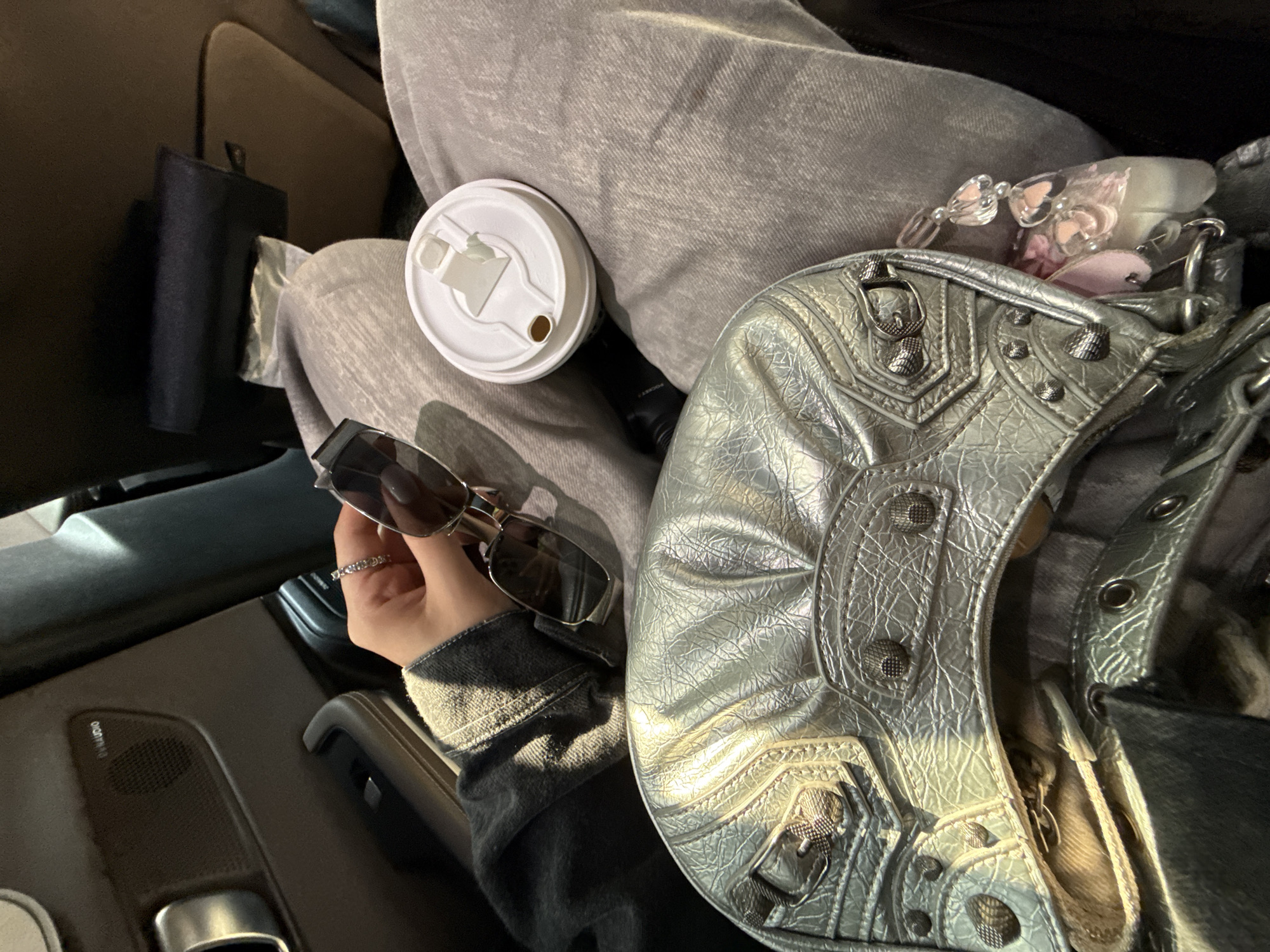 The image captures a close-up view inside a vehicle, likely a car, showing a person's lap, hand, and personal belongings. The scene is dominated by a pair of grey sweatpants or similar casual attire.
A hand, adorned with a ring on the finger, holds a pair of dark sunglasses, partially obscuring the view of the car's interior door panel and an armrest. On the person's lap, a white coffee cup with a lid is visible, positioned next to a silver, textured handbag. The handbag features numerous studs, buckles, and a distinct crinkled metallic finish, characteristic of a Balenciaga City bag style. Inside the bag, various small accessories are visible, including items with pink heart shapes.
In the background, behind the person's leg, a dark object resembling a wallet or small clutch can be seen. The car's interior is visible to the left, showing a dark-colored door panel with a speaker grille that has "PA audio" text printed on it. The lighting suggests daylight, with some sunlight reflecting off the metallic handbag, indicating it is likely daytime. The overall impression is of a passenger or driver on the move, carrying everyday items within a car. No specific activity beyond holding the sunglasses and having items on the lap is clearly depicted.