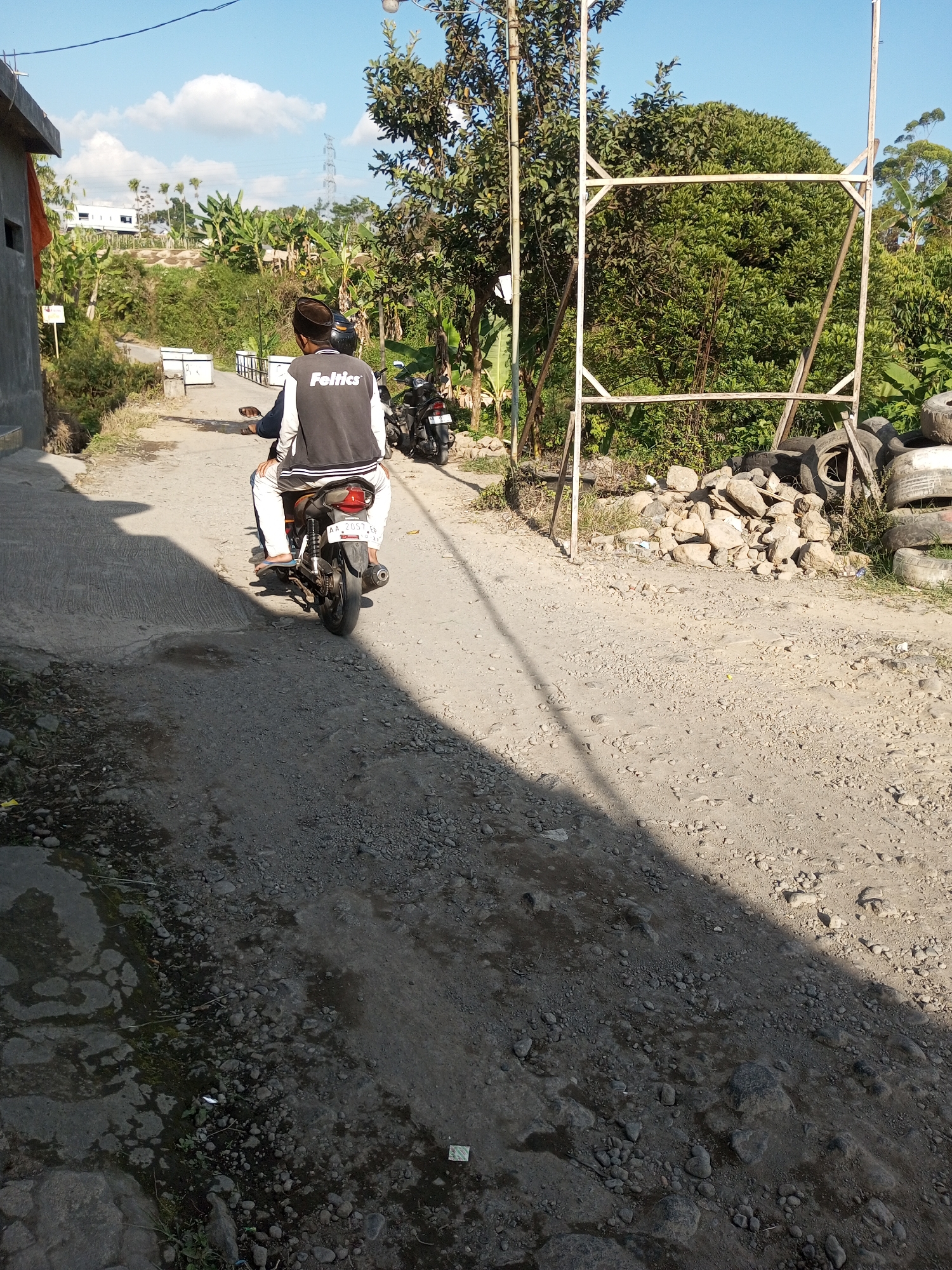 A rear view of two individuals on a motorcycle is captured on a paved road in Kalibebe, Indonesia. The driver, wearing a black and white jacket with "Feltics" emblazoned on the back, and a dark cap, is positioned in front. The passenger, wearing a white t-shirt, sits behind. The motorcycle is black and white, with a license plate reading "AA 2057 AA." The scene unfolds under a clear blue sky with scattered clouds. The road is flanked by lush green vegetation, including trees and banana plants. In the foreground, the road surface is a mix of asphalt and gravel, with a distinct shadow cast across it. To the right of the road, a pile of rocks and discarded tires are visible, next to a wooden frame structure. A building with a red tarp is on the left side of the road. A second motorcycle is parked further down the road on the right.