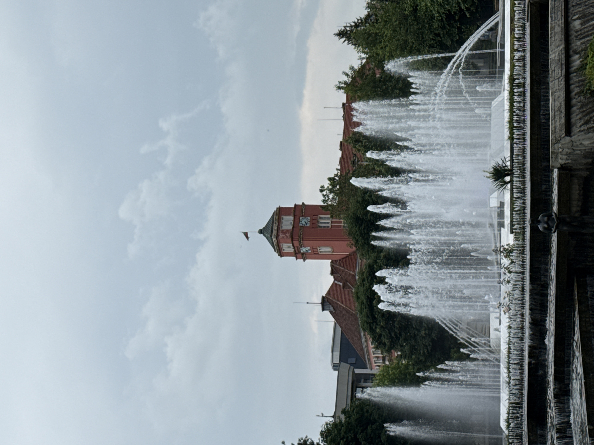 The image depicts a panoramic view of a multi-tiered fountain complex situated in the city of Pleven, Bulgaria, with architectural elements in the background under an overcast sky.
The sky occupies the upper third of the image and is uniformly gray with diffuse cloud formations. In the background, a prominent building with a red-tiled roof and a light red or pink facade is visible. This building features a distinctive clock tower topped with a greenish dome and a small flag, likely the Bulgarian national flag, fluttering. Multiple clock faces are visible on the tower. Surrounding this building and extending to both sides of the frame are dense clusters of green deciduous and coniferous trees. To the left of the main building, another structure with a flat, light-colored roof and dark walls is partially visible.
The midground is dominated by the elaborate fountain system. The uppermost tier of the fountain features numerous powerful vertical water jets, along with several arching jets creating dynamic water patterns. Below this, water cascades over multiple successive tiers, forming a stepped waterfall effect. The water is white and frothy as it falls. Green foliage, possibly low-lying shrubs or plants, is arranged along the edges of the fountain tiers.
In the foreground, more cascading water tiers are visible. On the lowest visible tier, towards the right side of the frame, a dark-colored statue of a nude human figure, likely female, stands partially submerged or at the edge of the water. The overall lighting is soft and diffused, consistent with the overcast conditions. There are no other human figures or active interactions explicitly visible.