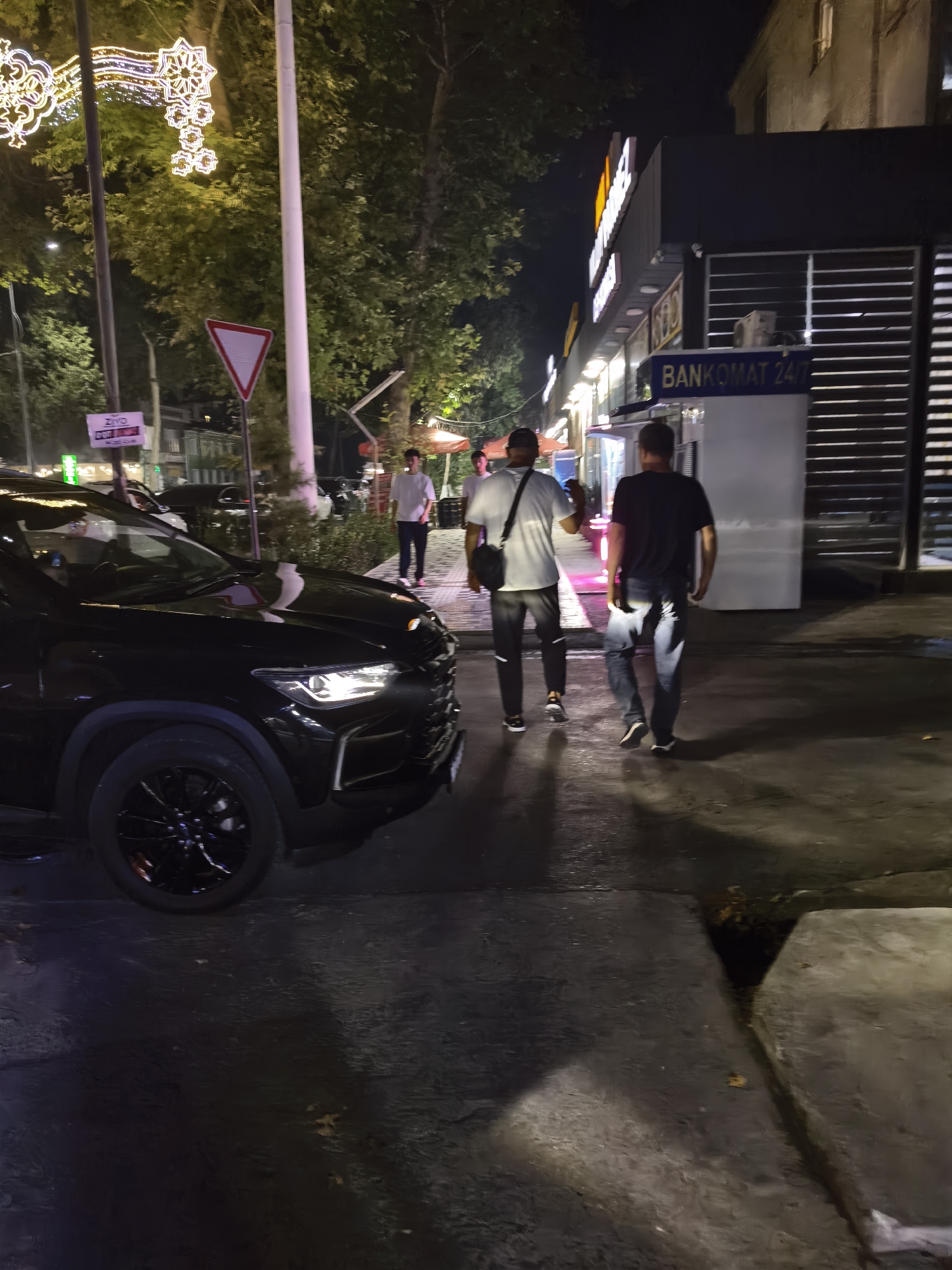 At night, on a street in Fergana City, Uzbekistan, two men are walking away from the camera. The man on the left is wearing a white t-shirt, dark pants with a white stripe, and black sneakers. He has a black bag slung across his body. The man on the right is wearing a black t-shirt, blue jeans, and black and white sneakers.
In the foreground, the front of a black SUV is visible, parked on the street. The SUV's headlights are on, illuminating the road. Behind the men, there is a building with a sign that says "BANKOMAT 24/7". There are also trees and streetlights visible in the background, suggesting an urban environment. Decorative lights, possibly for a holiday, are strung across the street in the upper left portion of the image. The ground appears wet, reflecting the streetlights and the lights from the building.