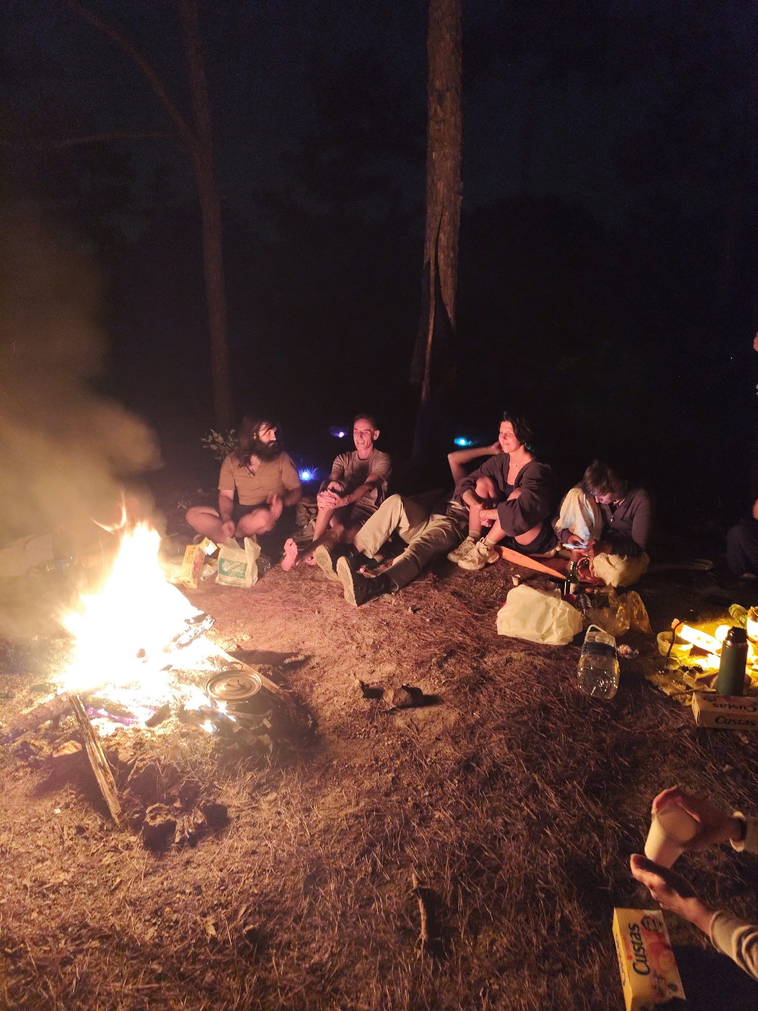 A group of approximately six people are gathered around a campfire at night. The location appears to be outdoors, possibly in a wooded area, as indicated by the presence of trees. The fire is burning brightly, casting light on the immediate surroundings and the people nearby. The individuals are sitting on the ground around the fire, and some appear to be interacting with each other, either talking or looking in each other's direction. There are food items scattered around them. The specific location within Lạc Dương, Vietnam, cannot be determined from the visual data alone.