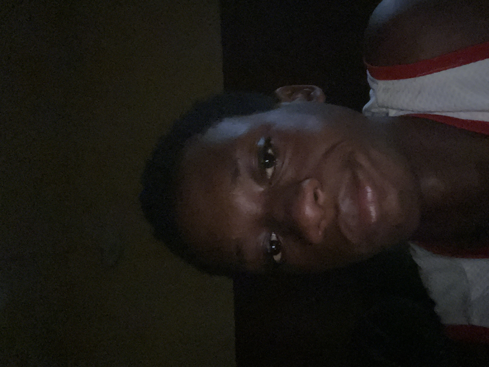 A dimly lit close-up of a dark-skinned individual with short, dark hair. The person is wearing a white tank top with red trim. The background is dark and indistinct. There are no clear indicators of the location being Nkpor, Nigeria.