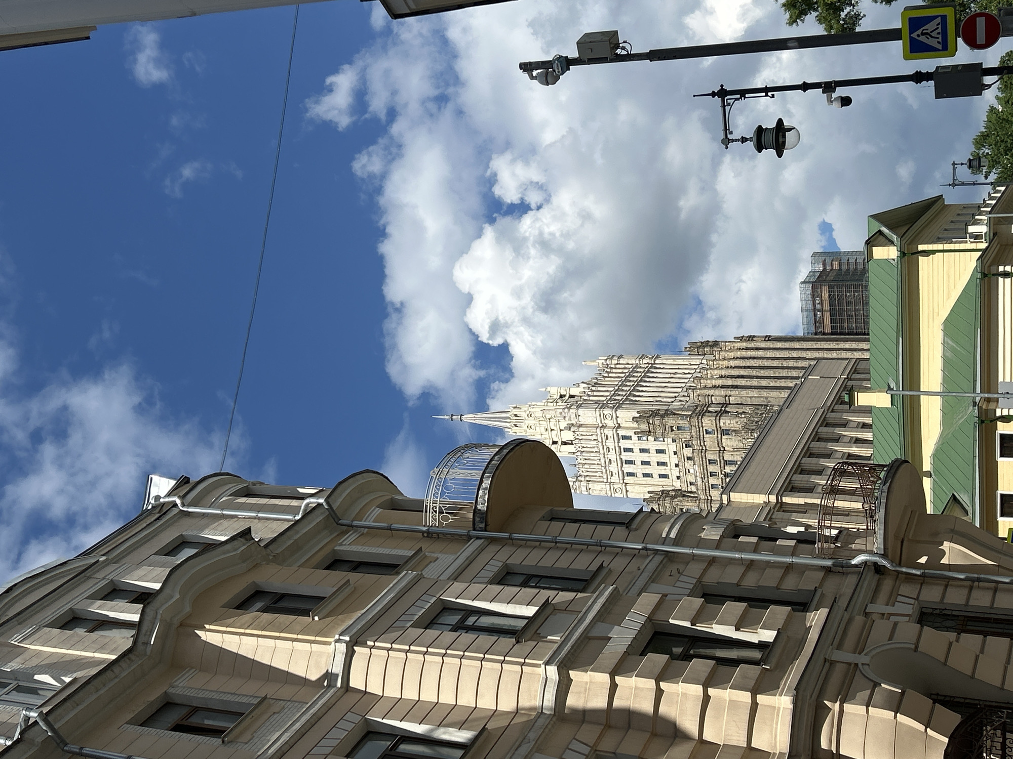 Captured from a low angle, the image showcases a variety of architectural elements juxtaposed against a vibrant sky. The focal point is a multi-story building in the foreground, characterized by its light-colored facade, distinct window placement, and ornate architectural detailing. Rising above this building is a taller, more elaborate structure, potentially a skyscraper or a significant civic building, rendered in white with a sharp spire that pierces the skyline. This structure appears older, with layers of setbacks, window patterns, and ornamentation. Adjacent to the primary building, there's another structure with a yellow and green exterior, possibly indicating a renovation or modern addition. Urban infrastructure is also visible, including a street lamp pole extending across the top right of the frame, complete with traffic signals and security cameras. The background features a striking blue sky filled with cumulus clouds, creating depth and contrast to the scene. Overall, the image appears to be taken in an urban setting in a city where historical architecture and modern urban infrastructure coexist.