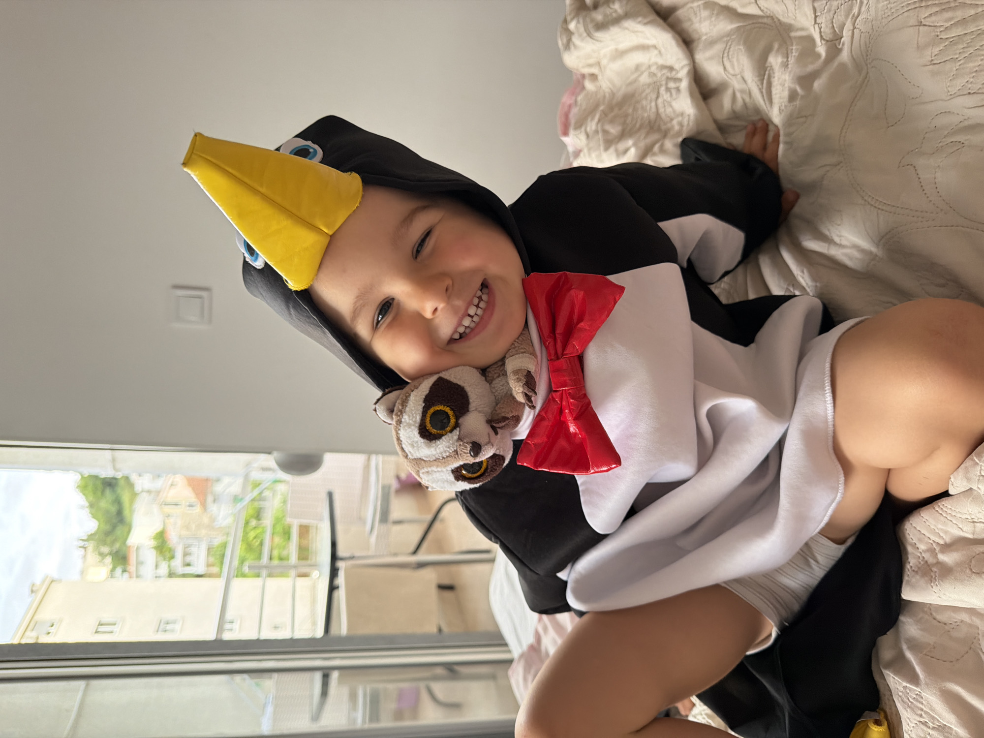 A child is lying on a bed indoors, wearing a penguin costume with a yellow beak and a red bow tie. The child is smiling and has a stuffed animal attached to the costume. The bed has a white, textured comforter. A window is visible in the background, showing buildings and greenery outside. Based on visual cues, the location is likely in Budva, Montenegro.