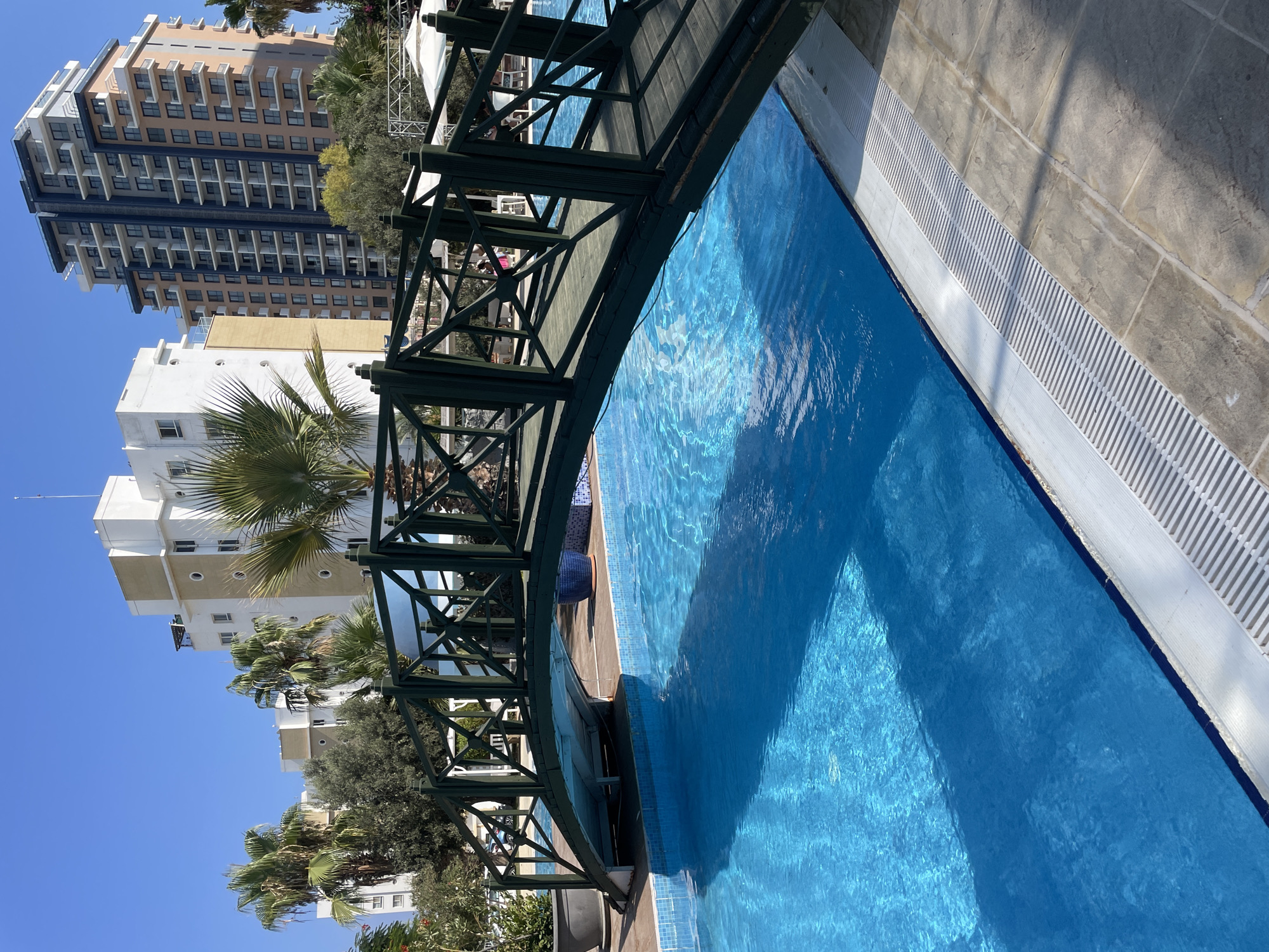 The image depicts a sunny outdoor scene, likely at a resort or hotel in Trikomo, Northern Cyprus. In the foreground, a clear blue swimming pool dominates the frame, with sunlight reflecting off its surface. A dark green wooden bridge arches over a section of the pool, its decorative lattice work visible. The pool area is bordered by tiled paving with a drainage grate. In the background, several multi-story buildings, some with modern architecture and balconies, are visible, interspersed with palm trees and other greenery. The sky is a clear, bright blue. There are no visible people in the immediate vicinity of the pool or bridge, but subtle hints of activity, such as chairs and umbrellas, can be seen in the distance near the buildings.