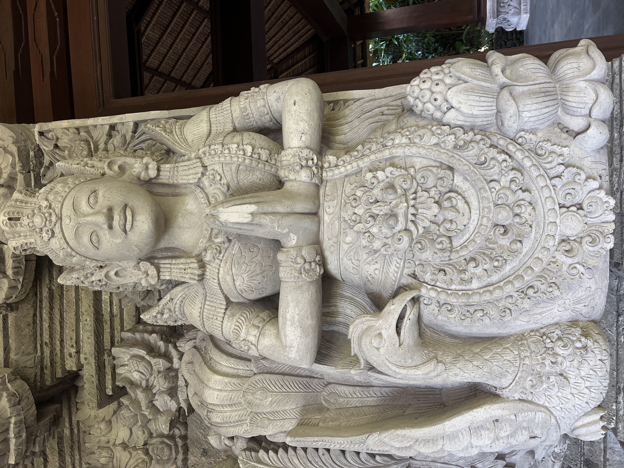 A carved, pale stone sculpture of a female figure is displayed in Kedewatan, Indonesia. The figure is adorned with intricate, circular designs on the garment that encircles her chest and shoulders. Her arms are bent at the elbow with her hands clasped together in front of her chest. Ornamental bracelets encircle her forearms. The lower portion of the sculpture extends to the left, carved with a bird-like form featuring feathers and a clawed foot. The sculpture is positioned near a structure with a dark brown, thatched roof visible in the background, along with wooden beams.