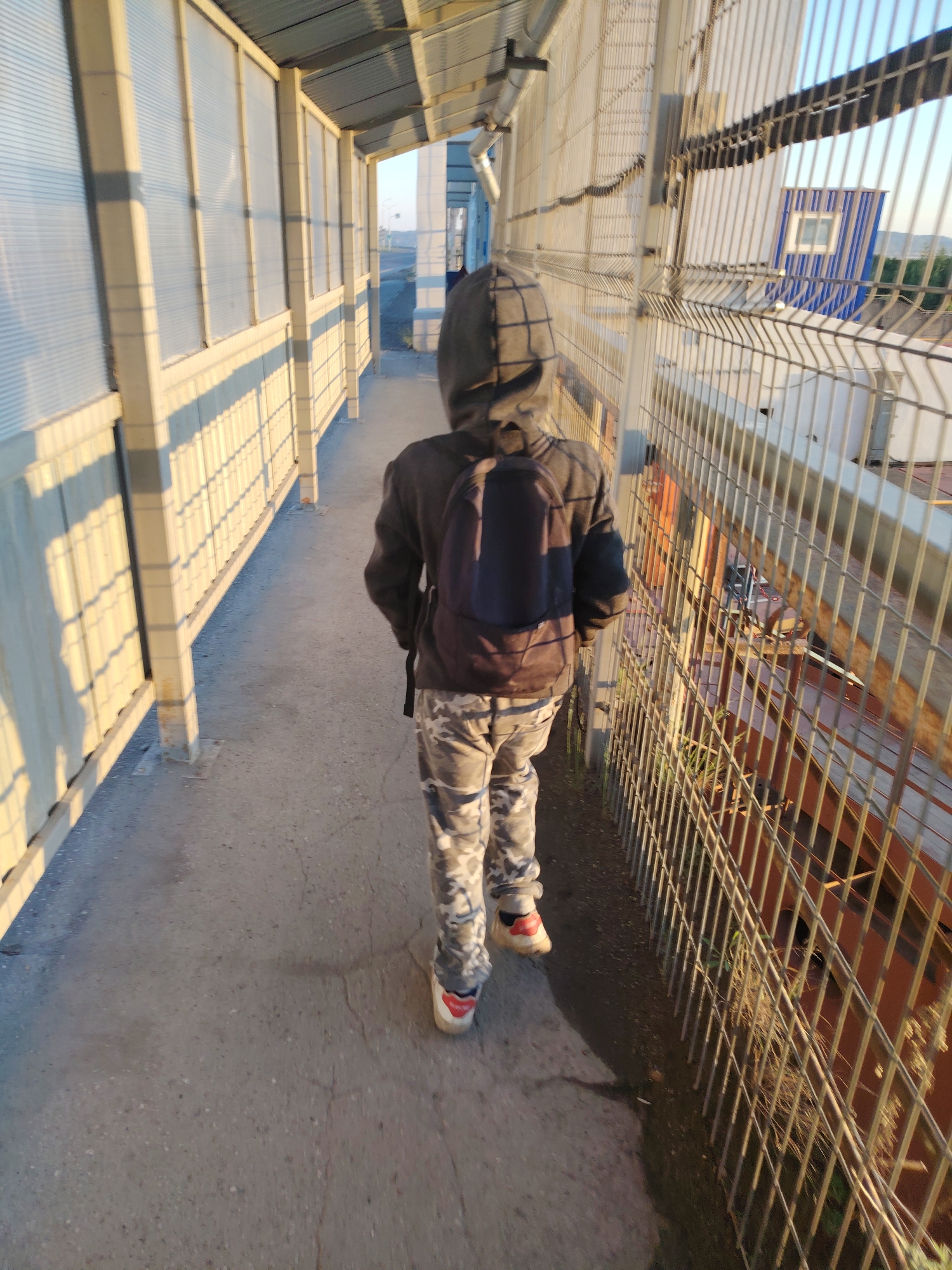 A person wearing a hooded jacket, camouflage pants, and sneakers walks down a covered walkway. The walkway has translucent panels on one side and a metal fence on the other. The sun casts long shadows. The scene appears to be outdoors, possibly in an urban or industrial area. The background shows a blue building with a white window, and some trees in the distance. The walkway is made of concrete, and there are metal supports holding up the roof.