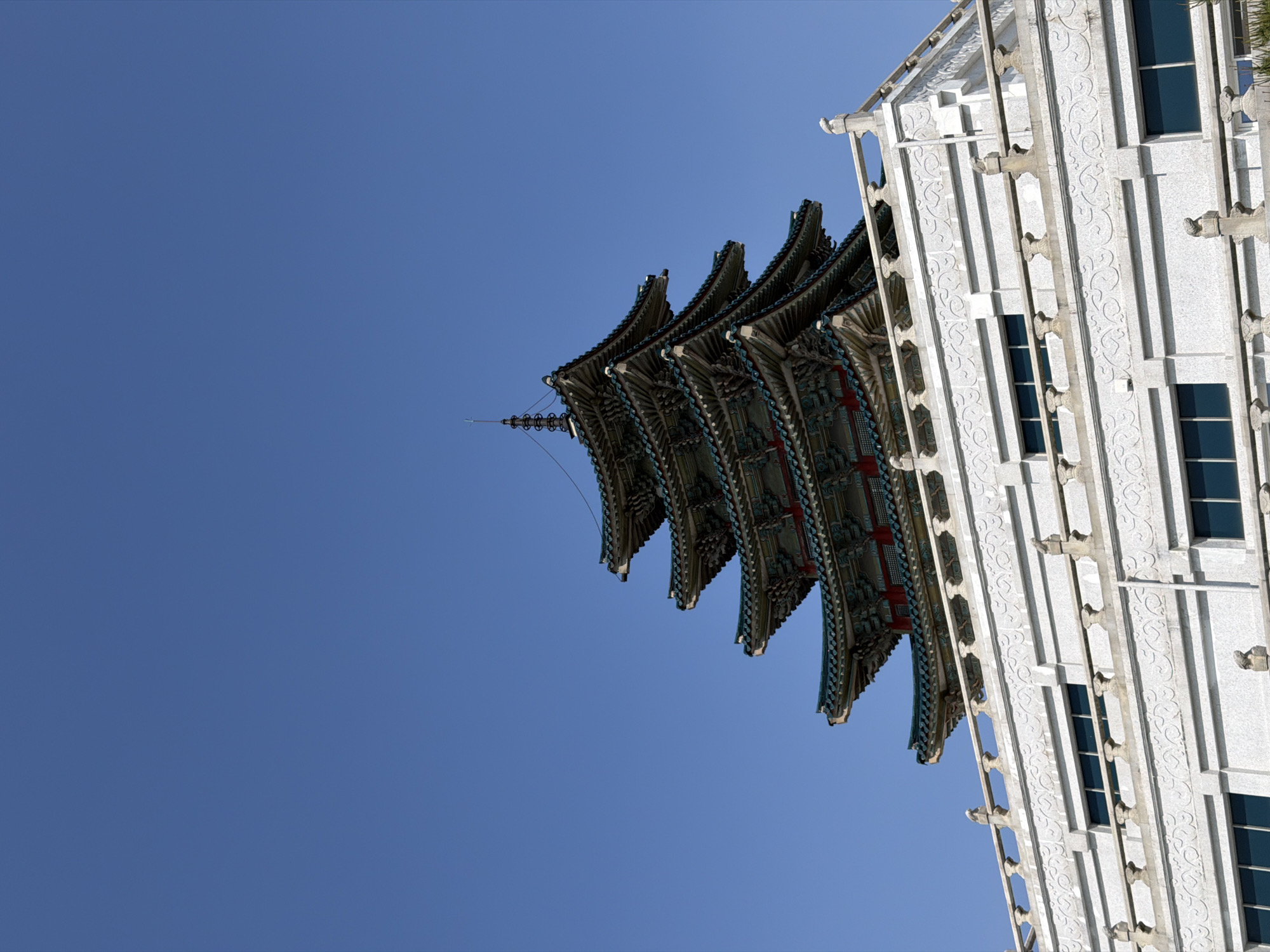 A low-angle shot shows the top of a traditional Korean building and a modern white building against a clear blue sky in Seoul, South Korea. The traditional building has multiple layered, ornate eaves with dark green roofs and colorful trim in blue, red, and white. The modern building is white with vertical columns, patterned facades, and rectangular blue windows. The structures are tilted, with the tops of the buildings appearing to converge towards the upper right corner of the frame.