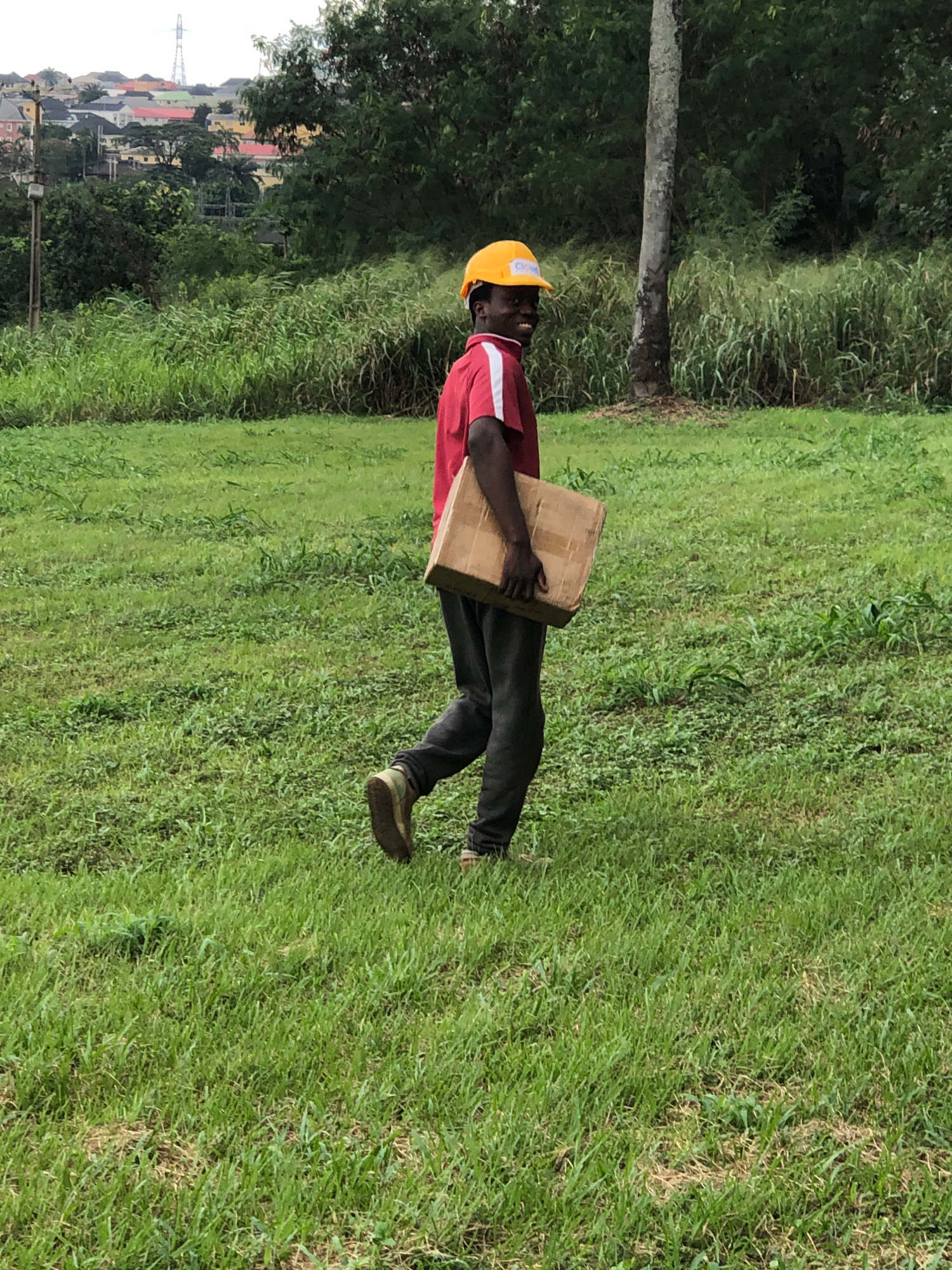 An individual wearing a red shirt and a yellow hard hat walks across a grassy field in Iju, Nigeria, carrying a brown cardboard box. The person is moving from left to right in the frame, with their head turned back to look towards the camera, smiling. Their posture suggests they are in motion. The background features lush green vegetation, a large tree, and a densely populated area with various colored buildings in the distance. The sky is overcast. The scene appears to be an outdoor area, possibly a construction site or a residential periphery.