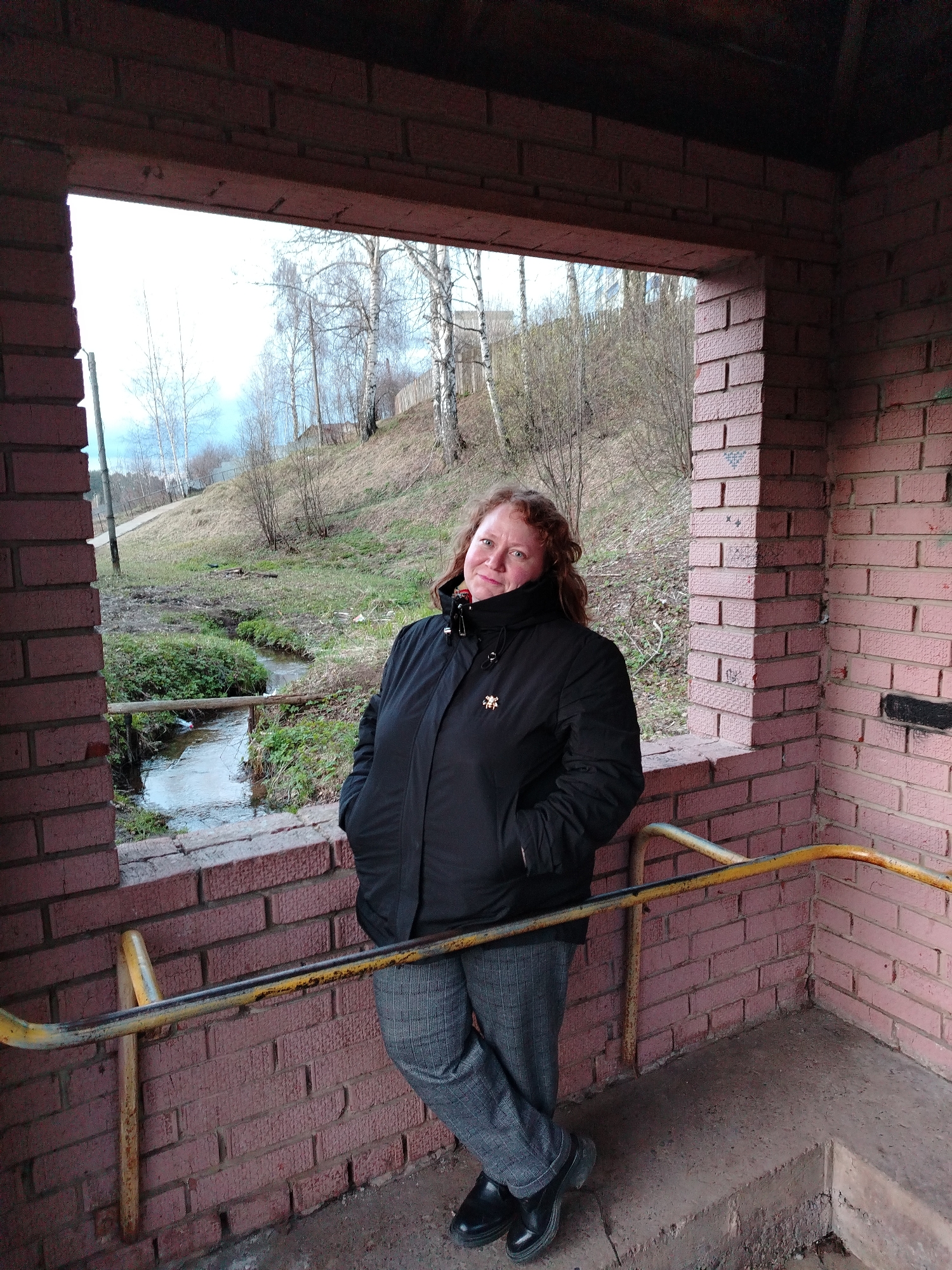 The image shows a middle-aged woman standing in a brick structure that overlooks a stream and a grassy hillside. The woman is wearing a black winter jacket, gray patterned pants, and black shoes. Her hands are in her pockets. The structure appears to be an open-air shelter, with brick walls and a metal railing. The surrounding environment includes a small stream, greenery, trees, and a hill. The sky is overcast. The location is Unknown, Unknown.