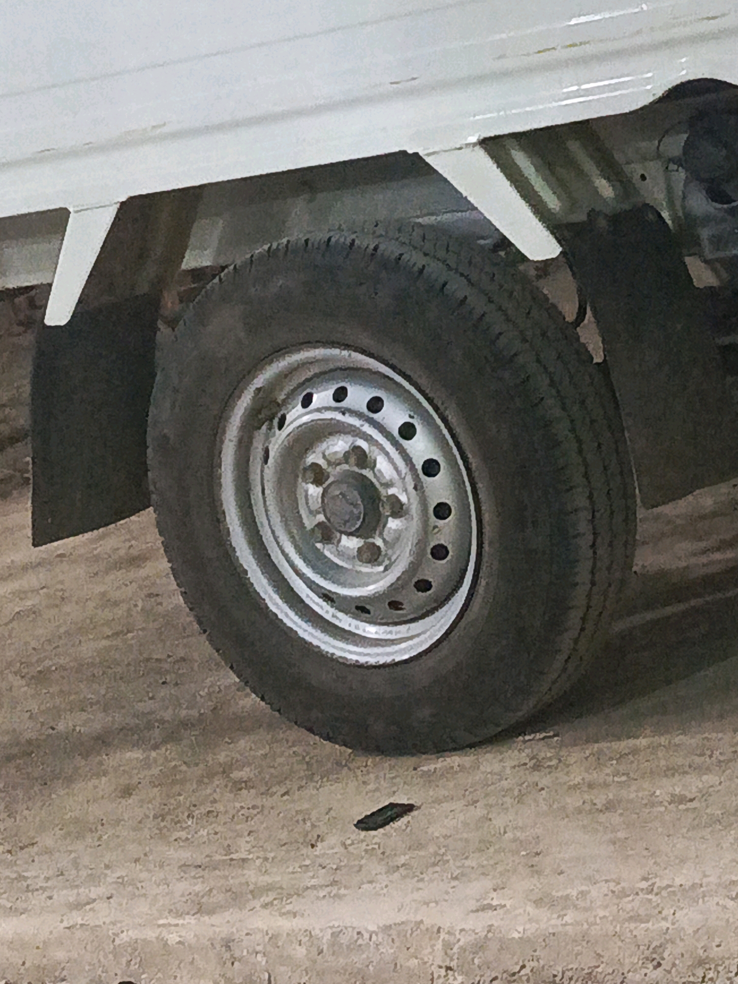 The image shows the rear wheel and undercarriage of a white utility vehicle parked on a dusty, light brown, uneven ground surface. The wheel has a silver rim with multiple circular holes. The tire is dark and appears to have some wear. A black mud flap is visible behind the wheel. The vehicle's chassis, painted white, is partially visible above the wheel. The environment suggests an outdoor setting, possibly a rural or unpaved area. There are no people or specific landmarks visible that would indicate the exact location in Megang Sakti V, Indonesia.