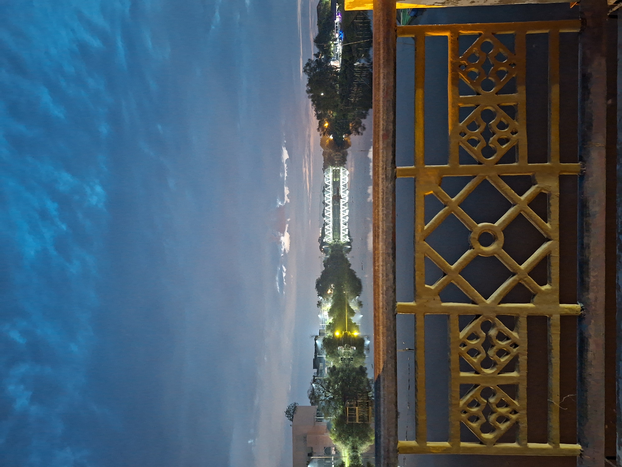 This image depicts a twilight view of the Krueng Cunda river in Lhokseumawe, Indonesia, seen from a bridge railing. The sky is a deep blue with scattered clouds, reflecting on the calm water. On the far bank, buildings, trees, and streetlights are visible, their lights creating a warm glow against the darkening sky. A bridge, illuminated with rows of lights, spans the river in the distance. The foreground is dominated by the weathered yellow railing of the bridge, featuring a decorative geometric pattern. The scene is devoid of any people, conveying a sense of quiet evening ambiance.